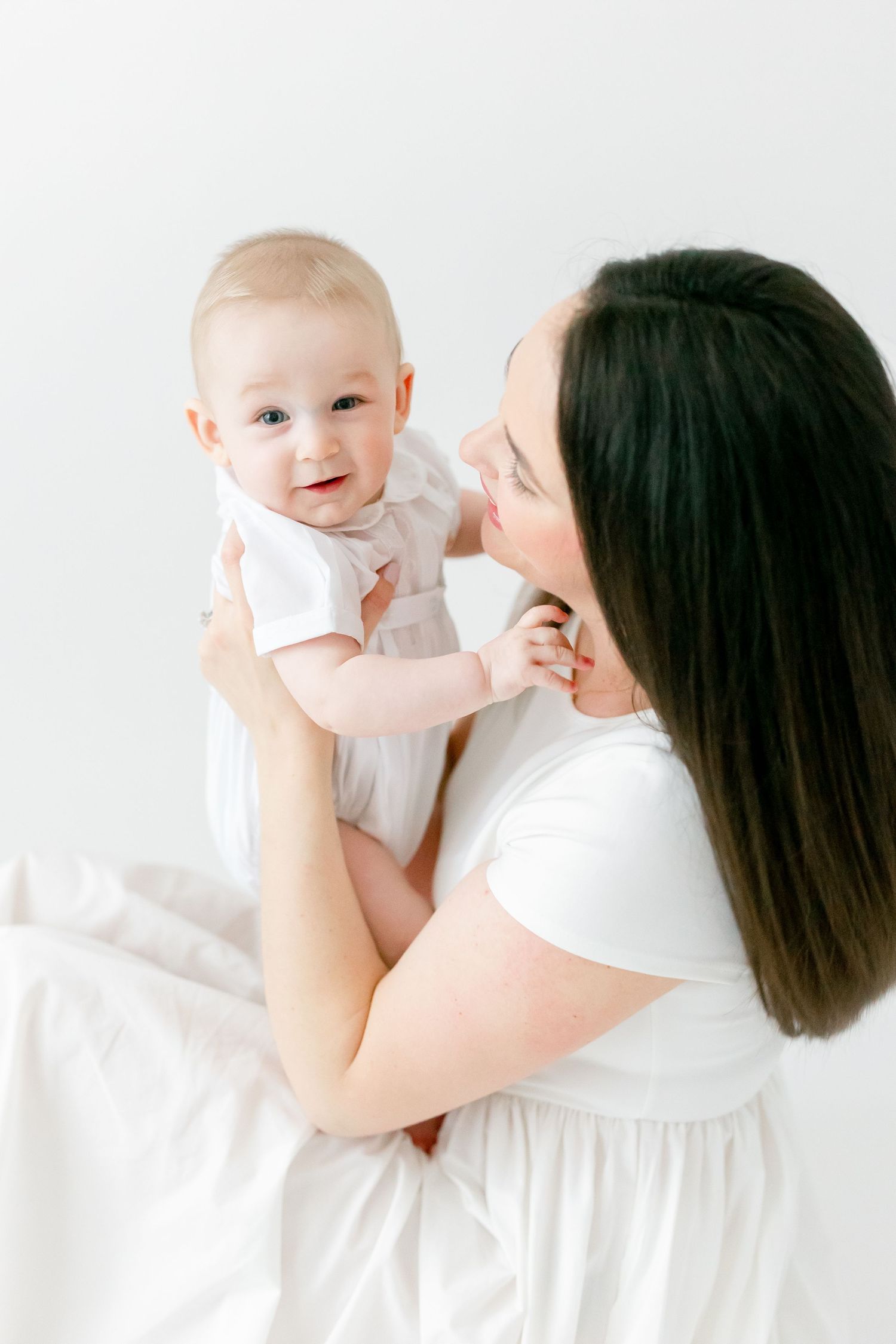 A smiling baby in a white outfit is held up by a dark-haired mother in a white dress against a bright, clean white studio background.