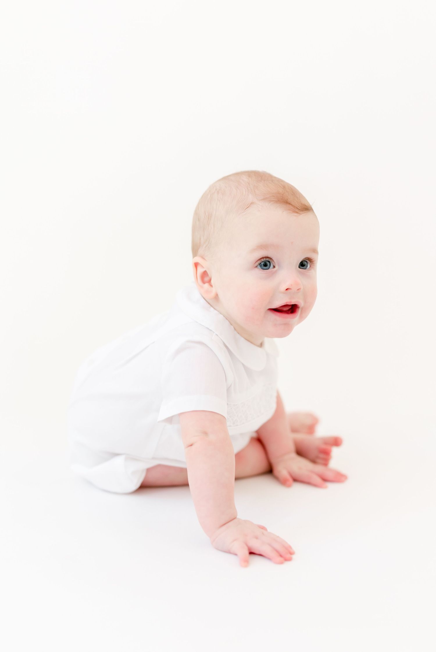 Smiling baby with blue eyes in a white collared romper crawling on a clean white studio background.