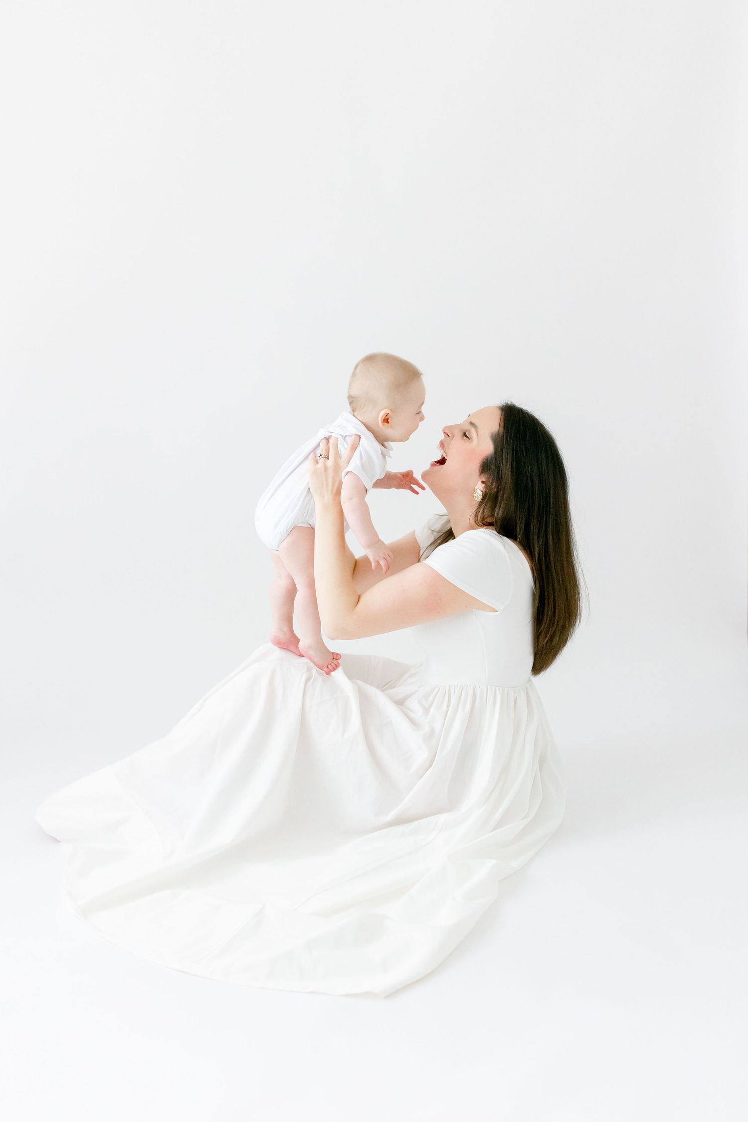 A mother in a white dress sits on a white floor, joyfully lifting her baby while smiling and playing together.