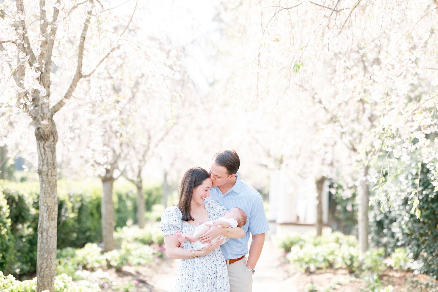 A couple shares a tender moment in a sunlit garden with blooming trees and greenery in the background.