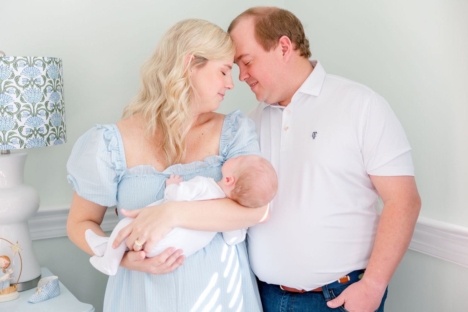 New parents tenderly touch foreheads while mother holds sleeping newborn baby in a softly lit nursery.