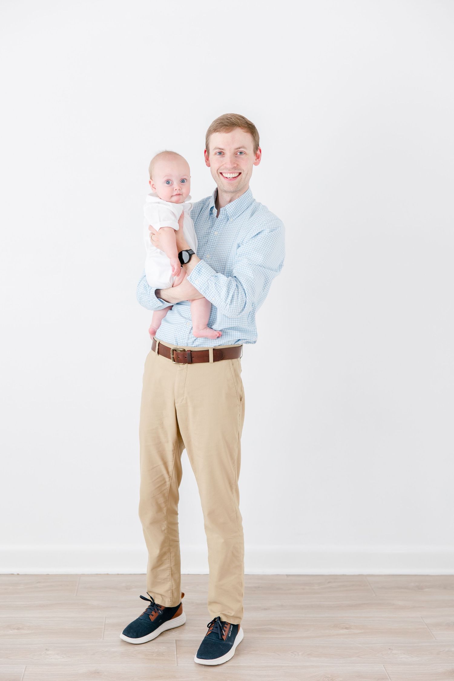 Father in light blue shirt and khakis holds baby in white onesie against white backdrop.