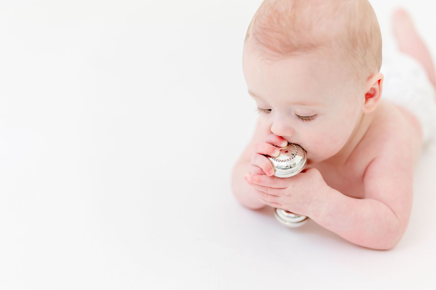 Baby lying on its tummy against a white background while chewing on a small silver rattle held in both hands.