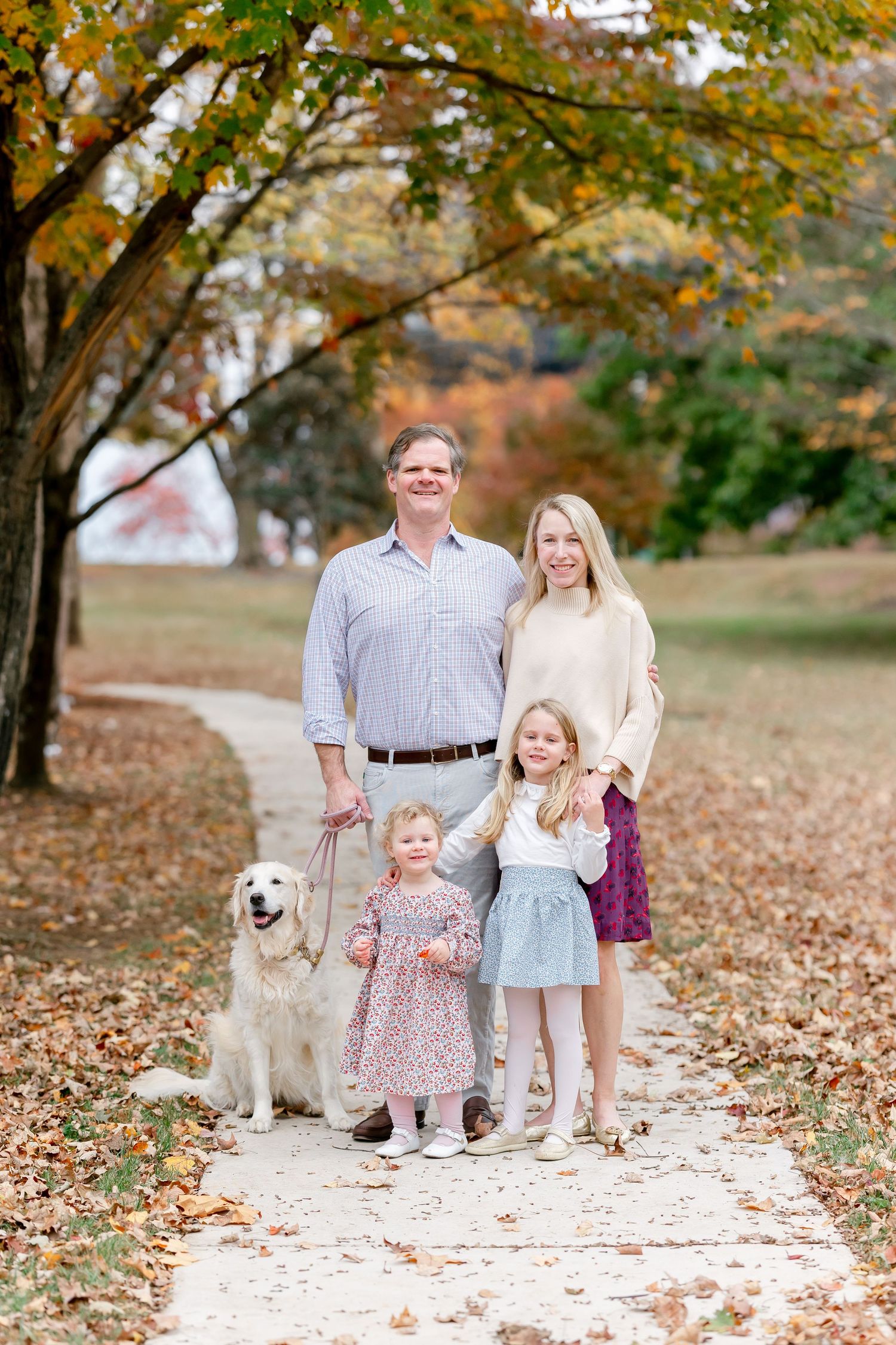 Family of four with golden retriever poses on leaf-covered path surrounded by colorful autumn foliage.