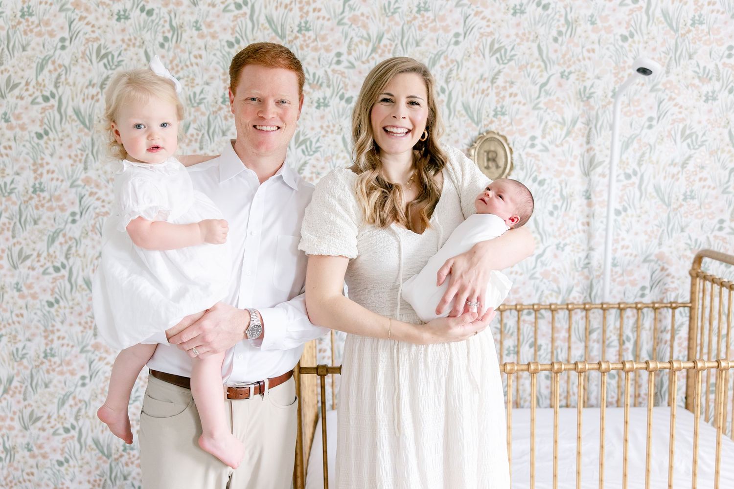 Family of four wearing white outfits pose together against floral wallpaper in a bright room with rattan furniture.