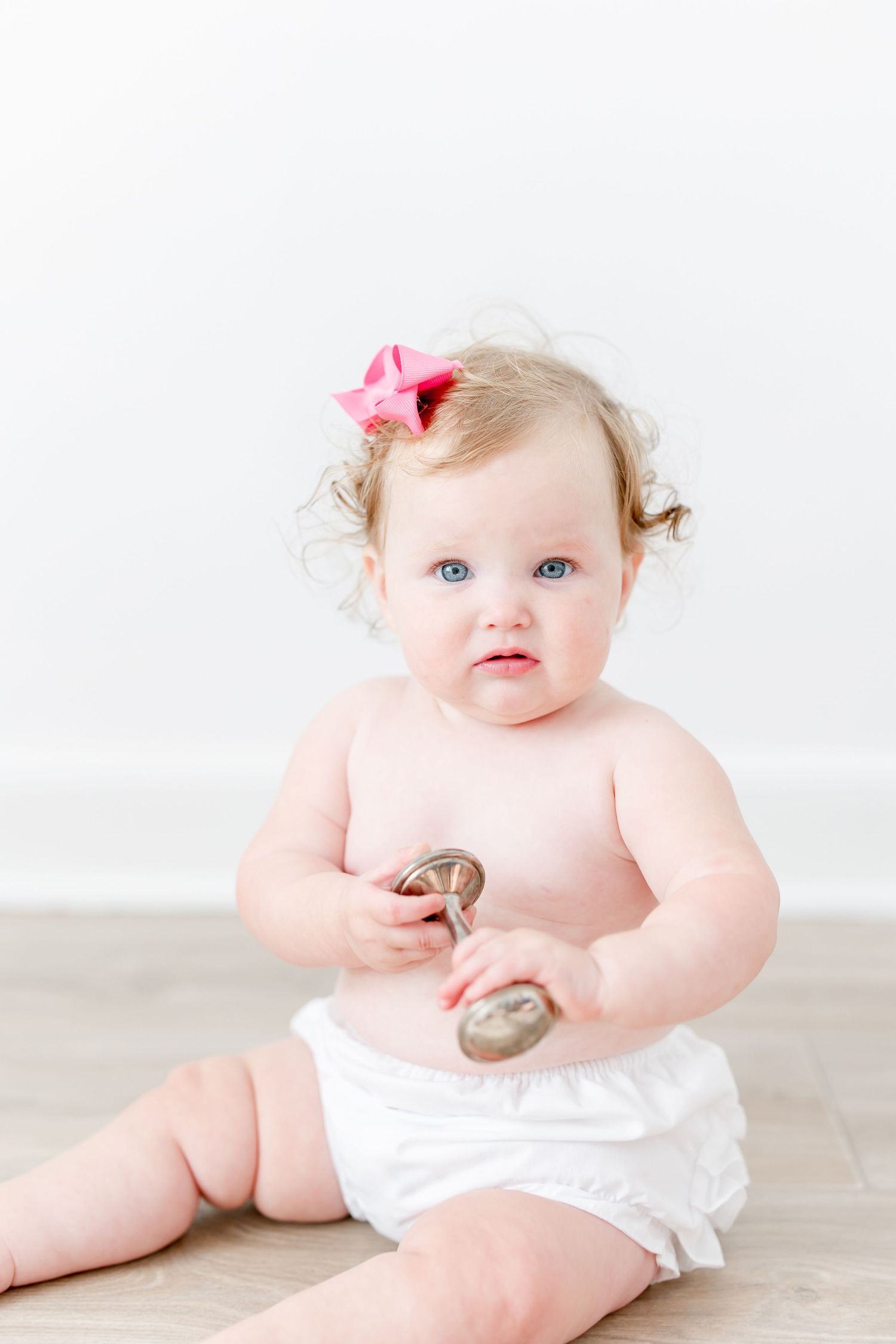 Sweet baby in white bloomers sits on wooden floor holding rattles with pink bow in curly hair.