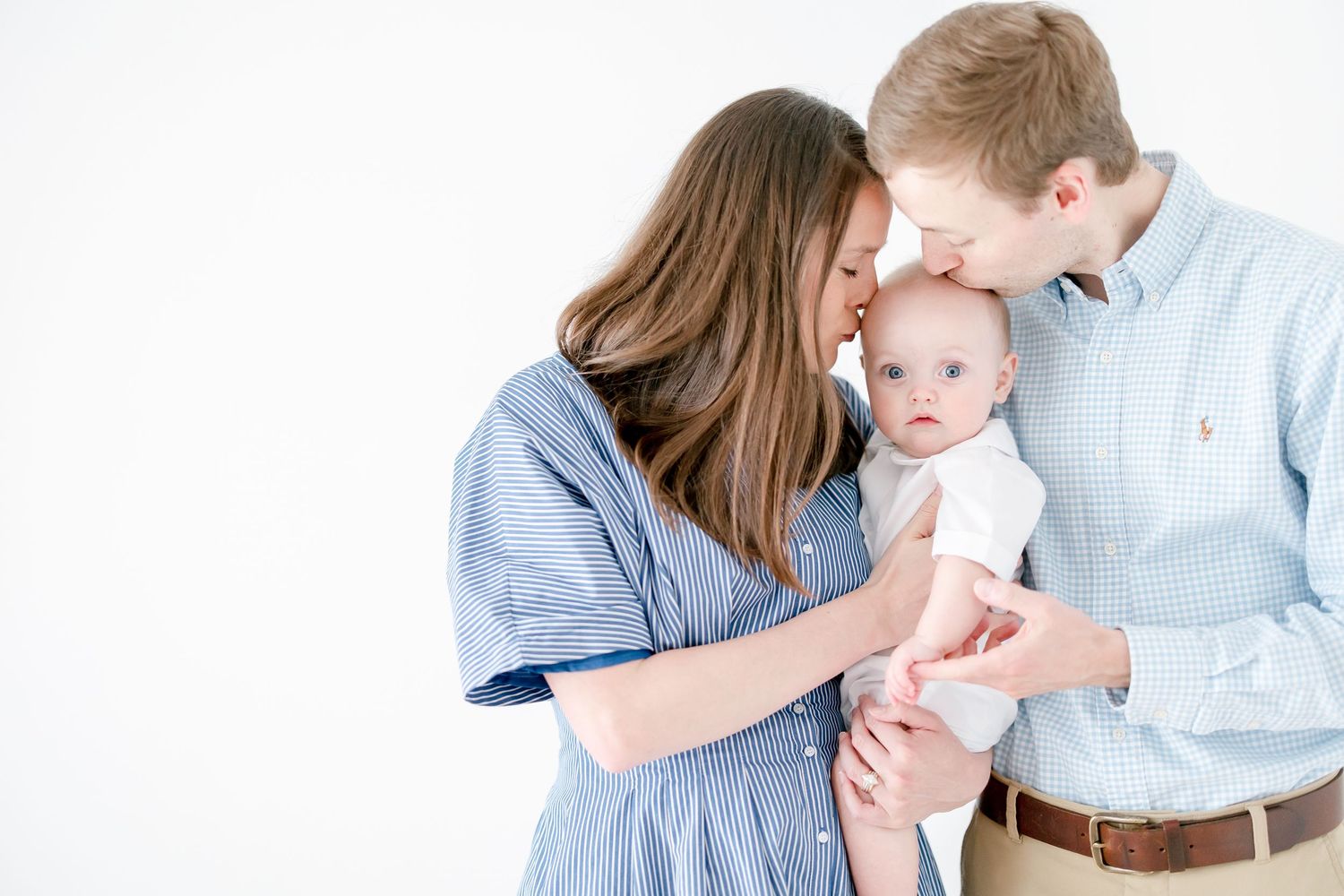 Parents in blue clothing share a tender moment with their baby against a white background.