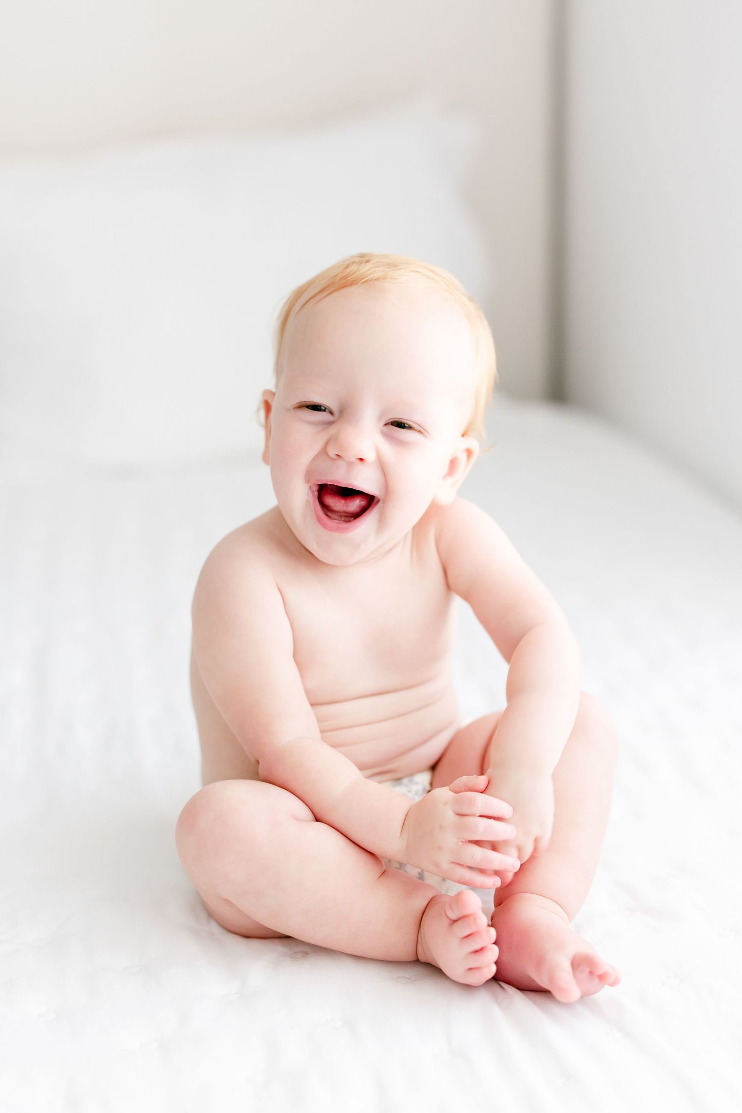 A laughing baby sits on white bedding wearing only a diaper while holding their foot.