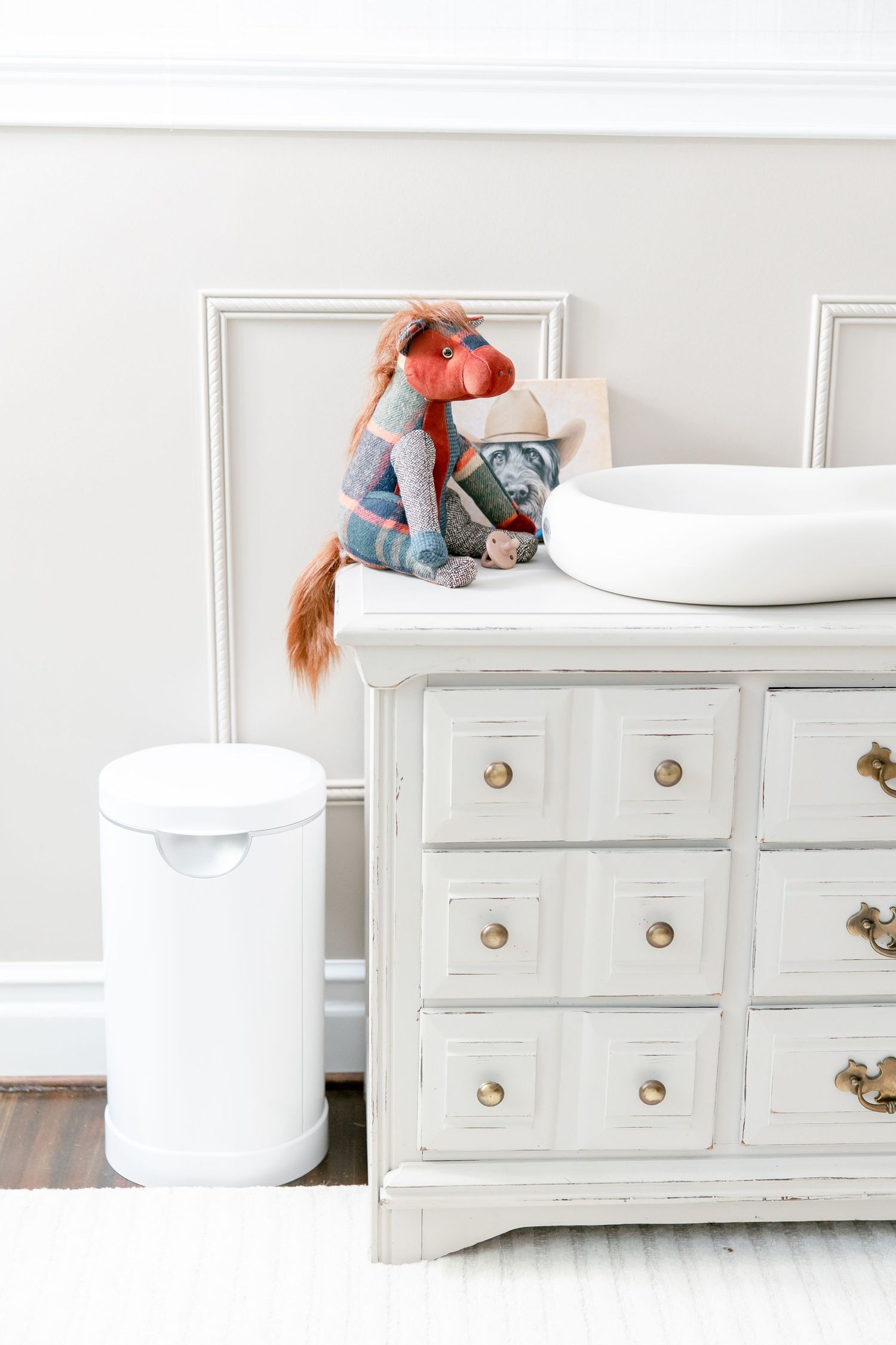 A decorative white dresser with brass handles displays a plush fox toy and pillow next to a white diaper pail.
