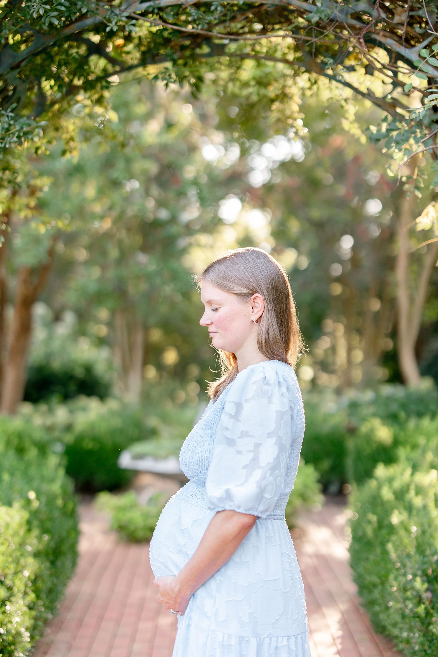 Woman in flowing white dress poses along brick pathway in garden setting.