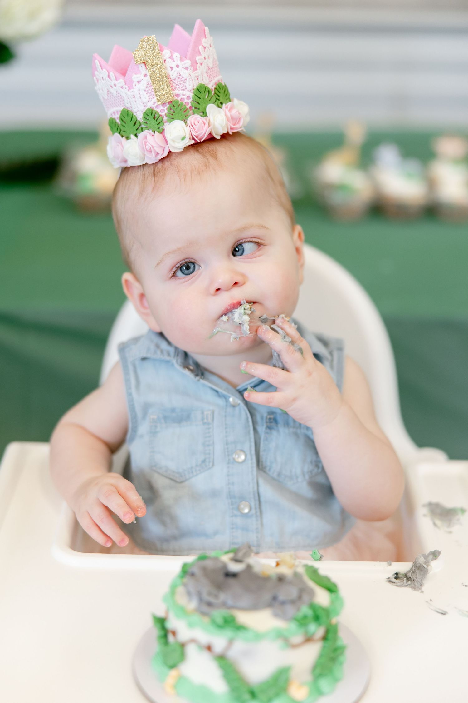 Baby wearing pink and green crown while eating birthday cake in high chair.