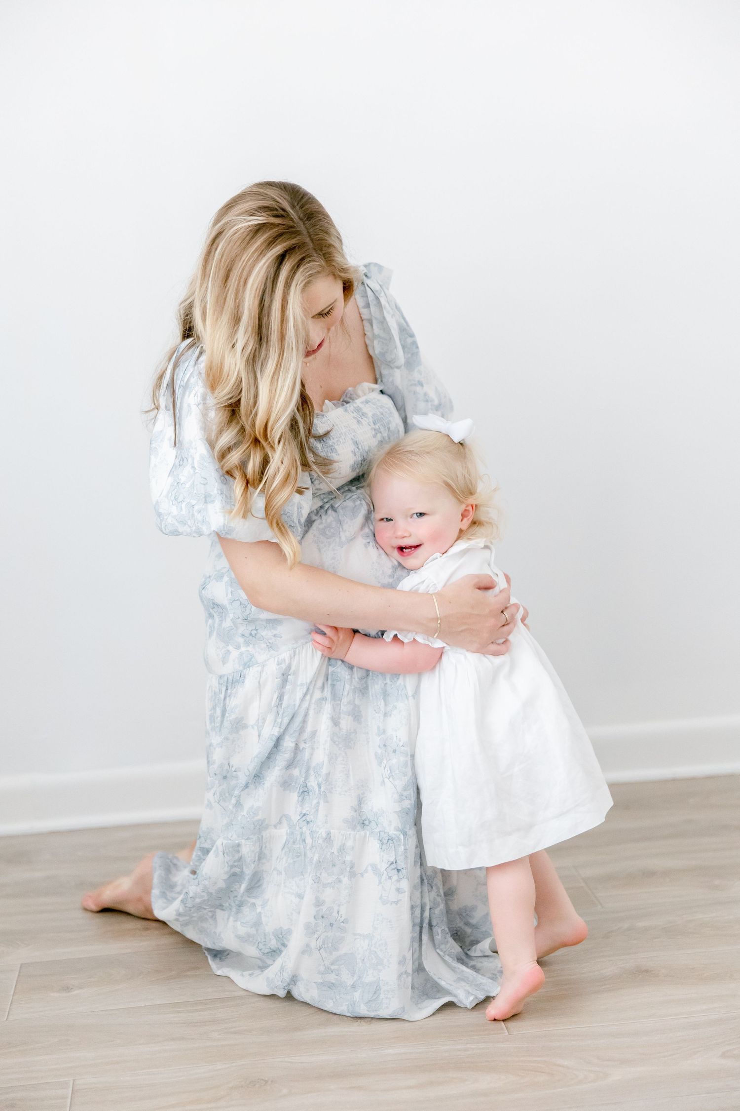 A tender moment between parent and child as they embrace, wearing a blue floral dress and white dress on wood floors.