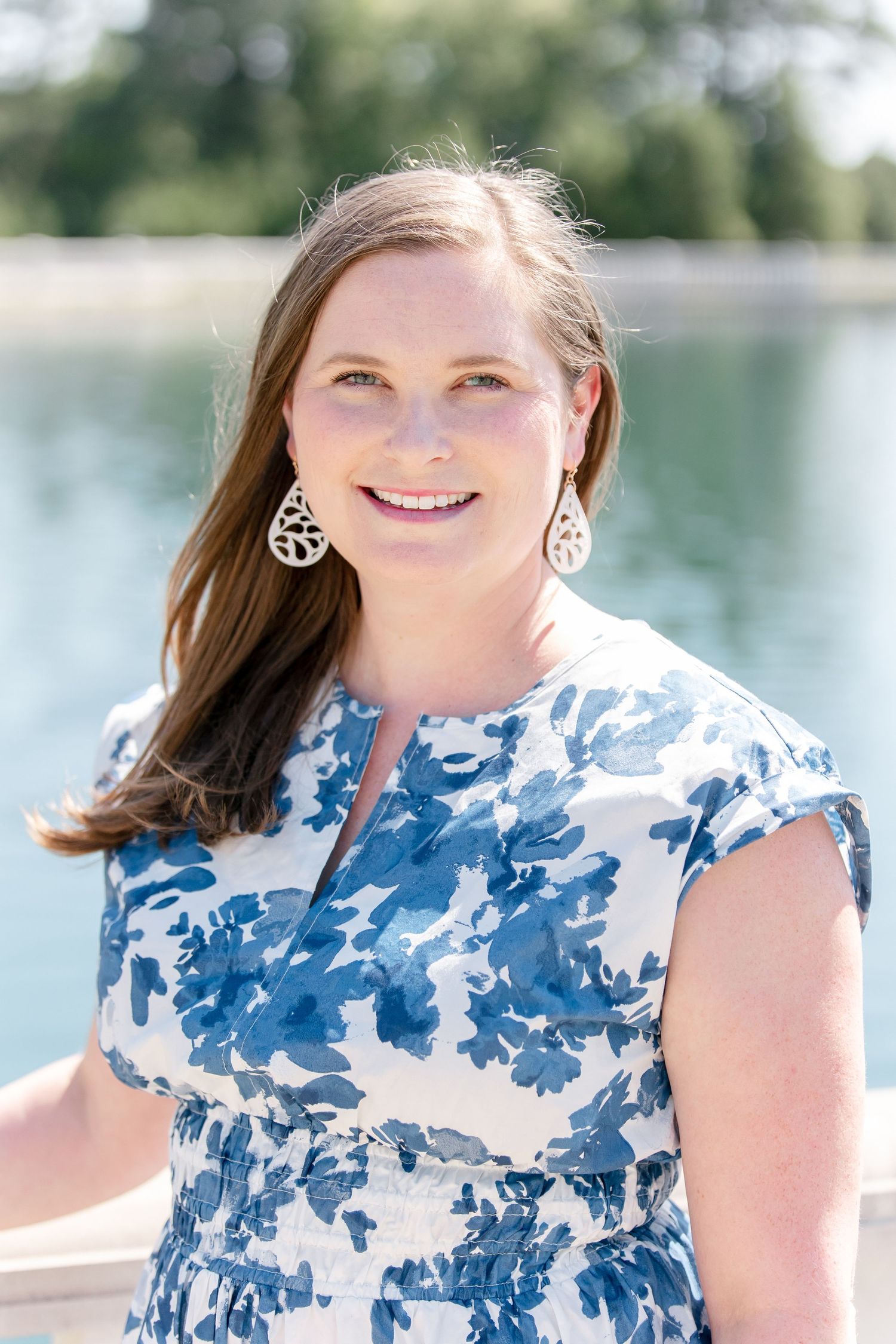 A smiling portrait in a blue and white floral dress wearing decorative earrings by a waterfront.
