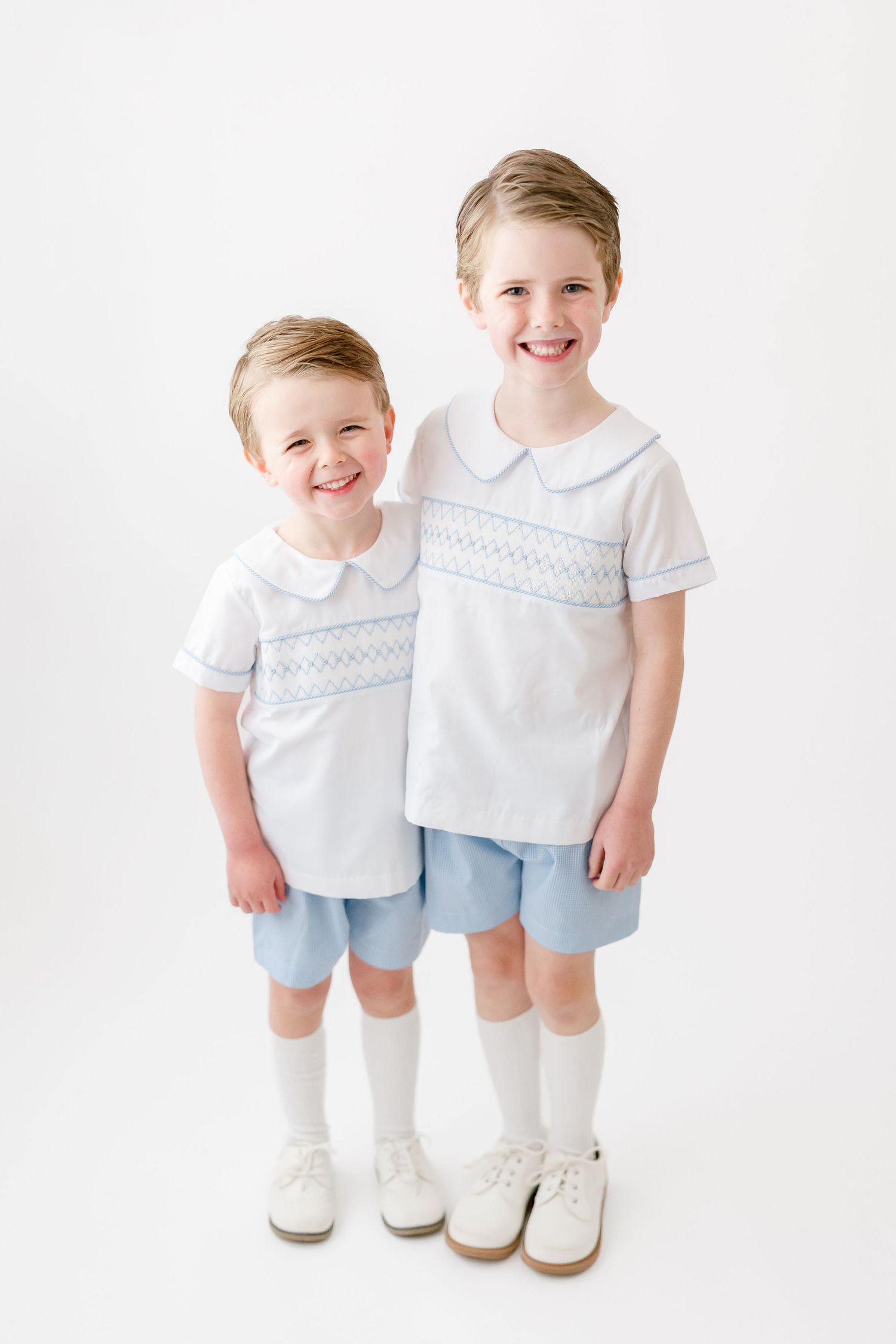 Two children wearing matching white smocked shirts and light blue shorts with white knee socks and shoes against white background.