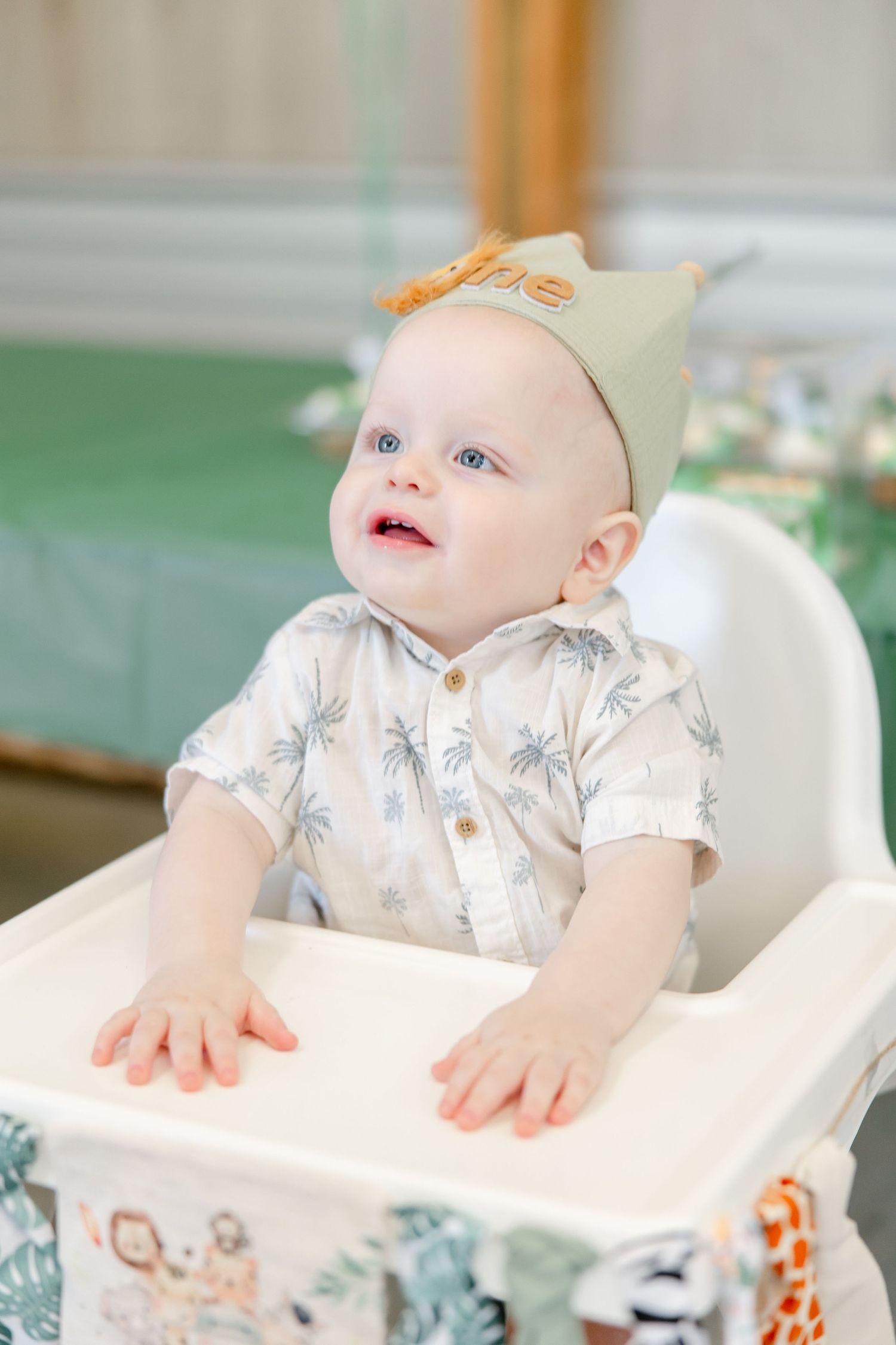 Baby in highchair wearing beige crown and palm print shirt at party setting.