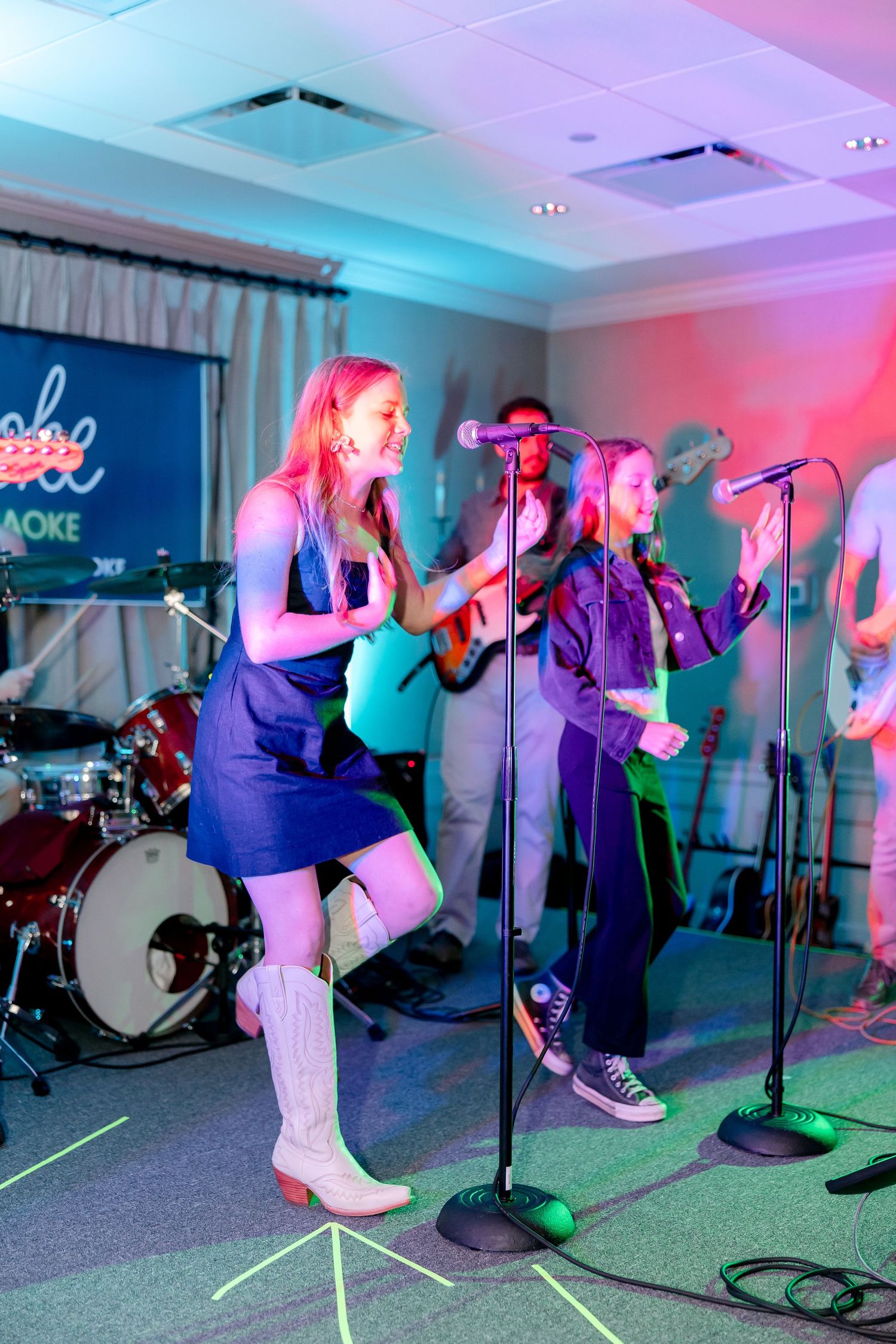 Female performers singing on stage with colorful lighting and musical equipment in background.