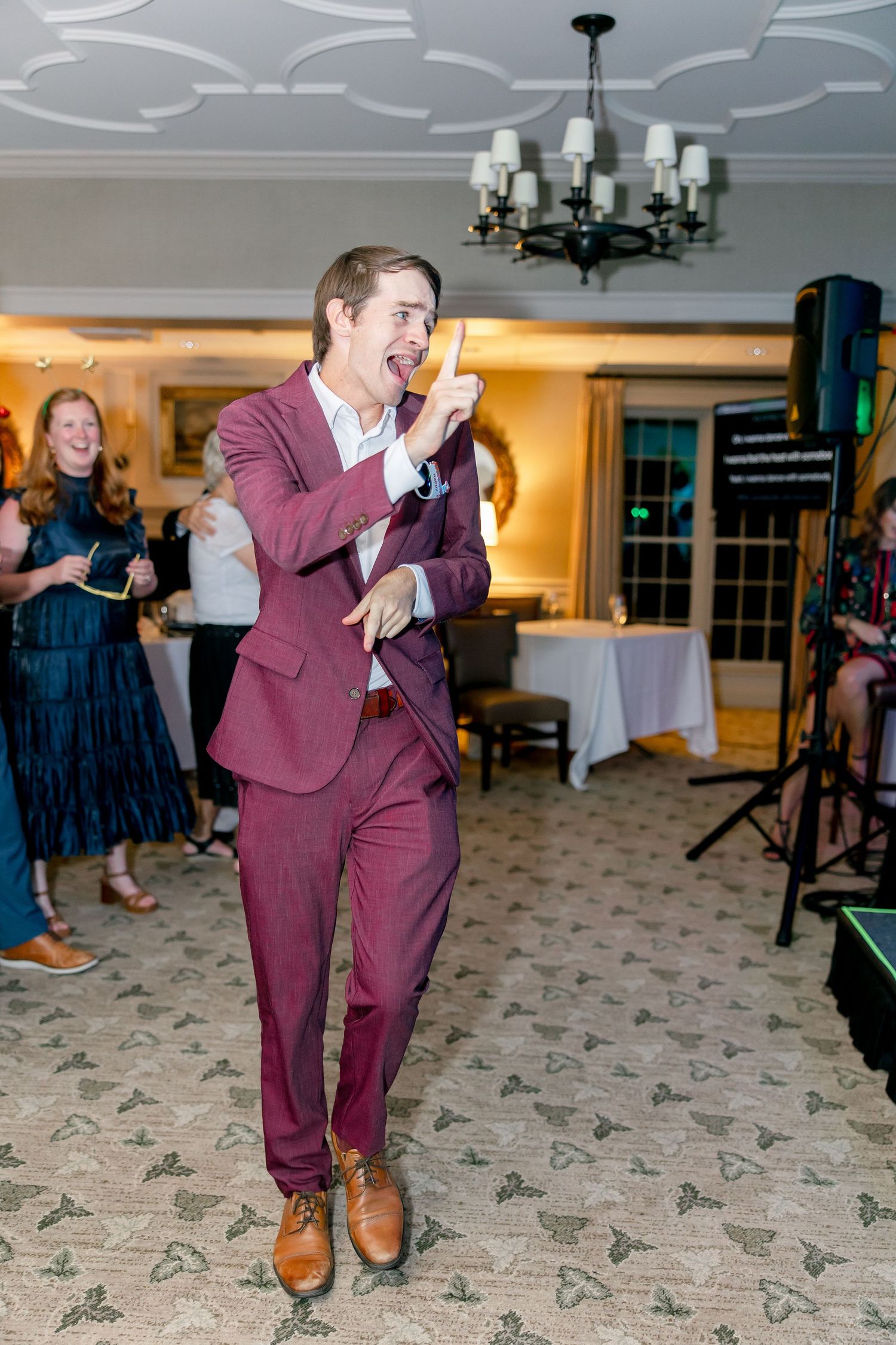 Enthusiastic person in burgundy suit singing or speaking into microphone at formal event with chandelier overhead.