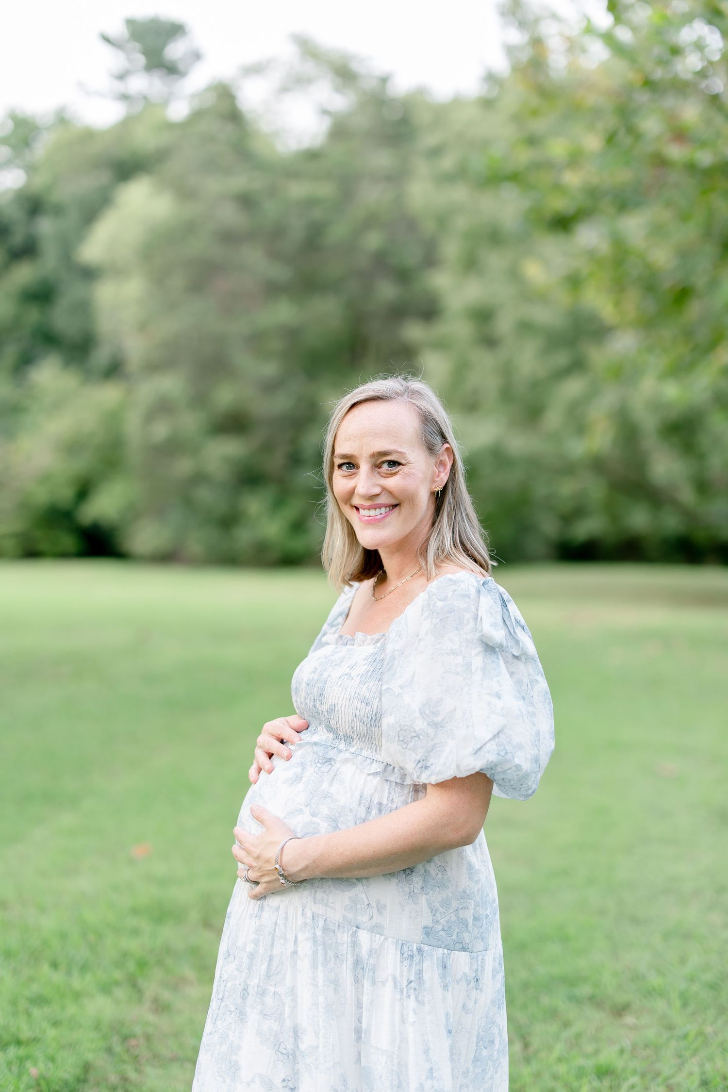 Woman in light blue floral dress stands smiling in a serene park setting.