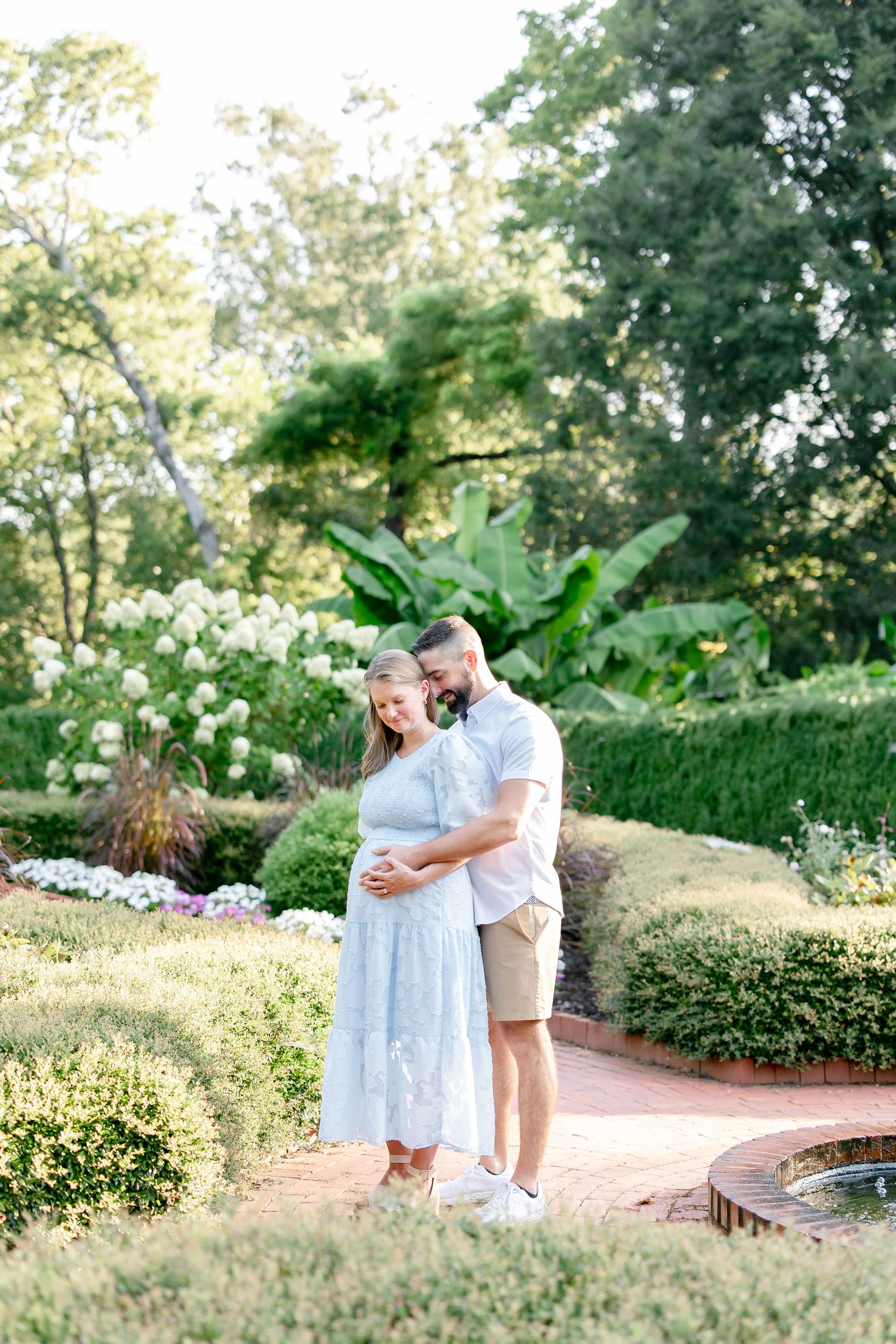 A couple embraces in a romantic moment on a brick pathway surrounded by lush garden landscaping in soft sunlight.