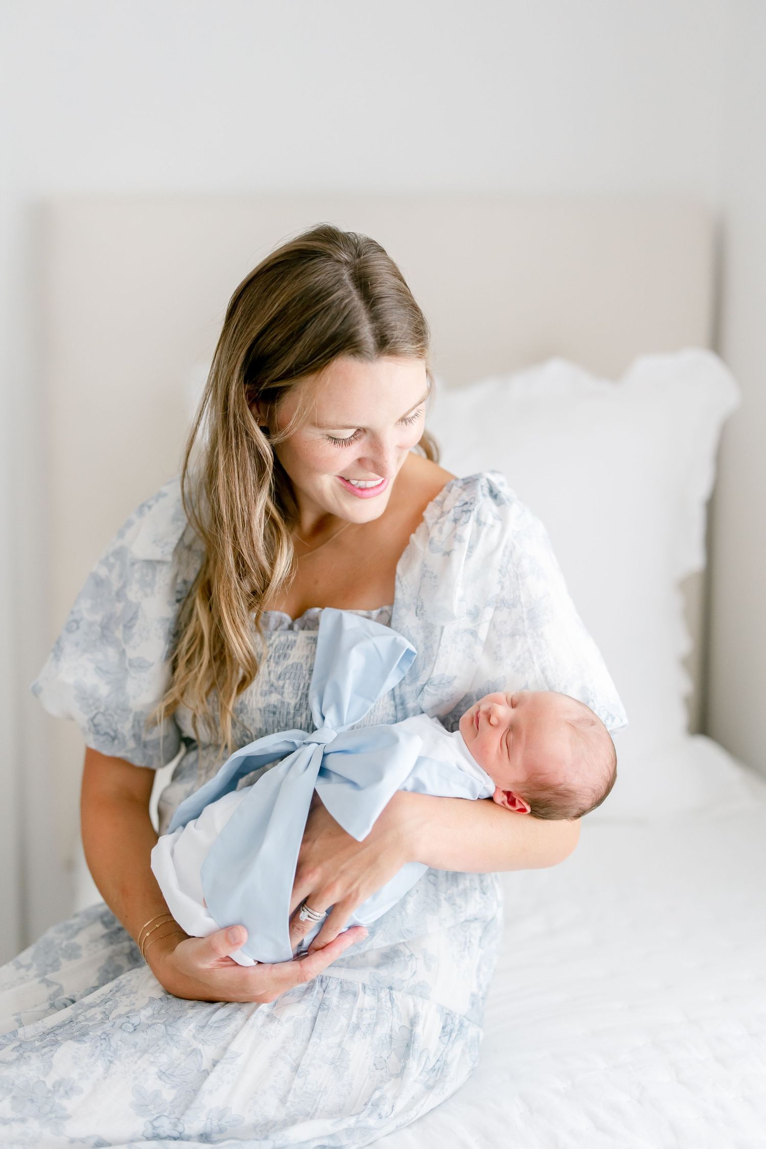 Loving parent in soft blue dress cradles sleeping newborn baby on white bed in a series of gentle poses.