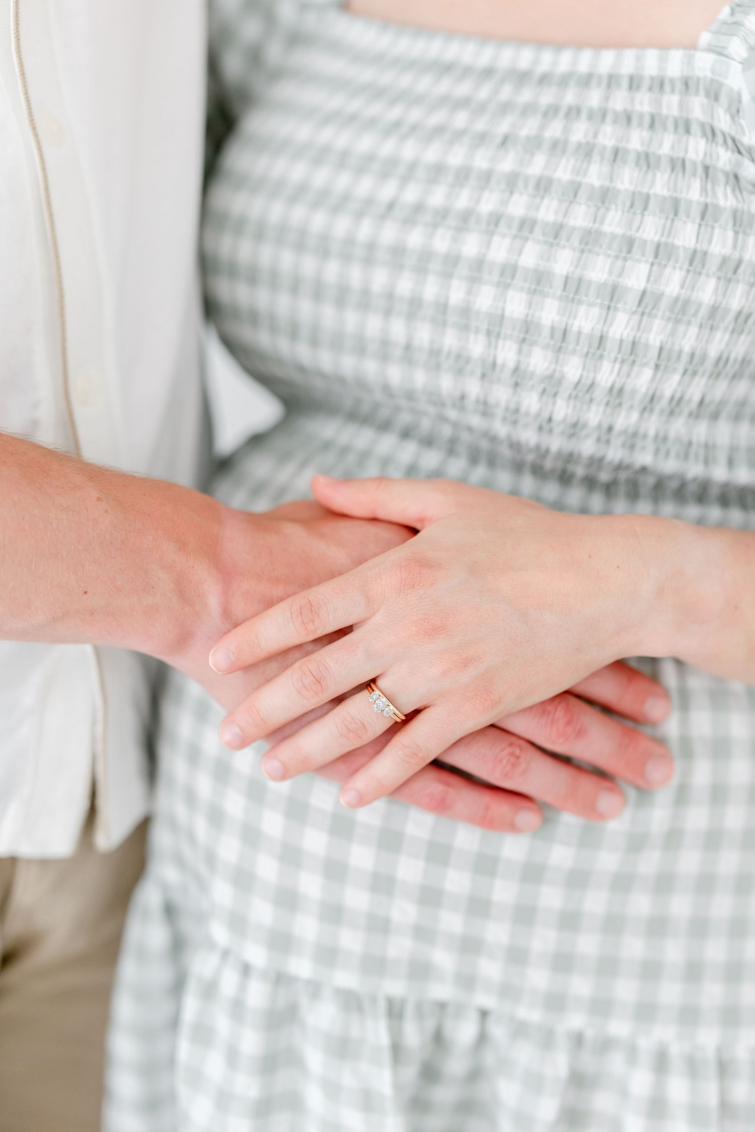 Hands with engagement ring shown against a light blue and white gingham dress in a tender moment.