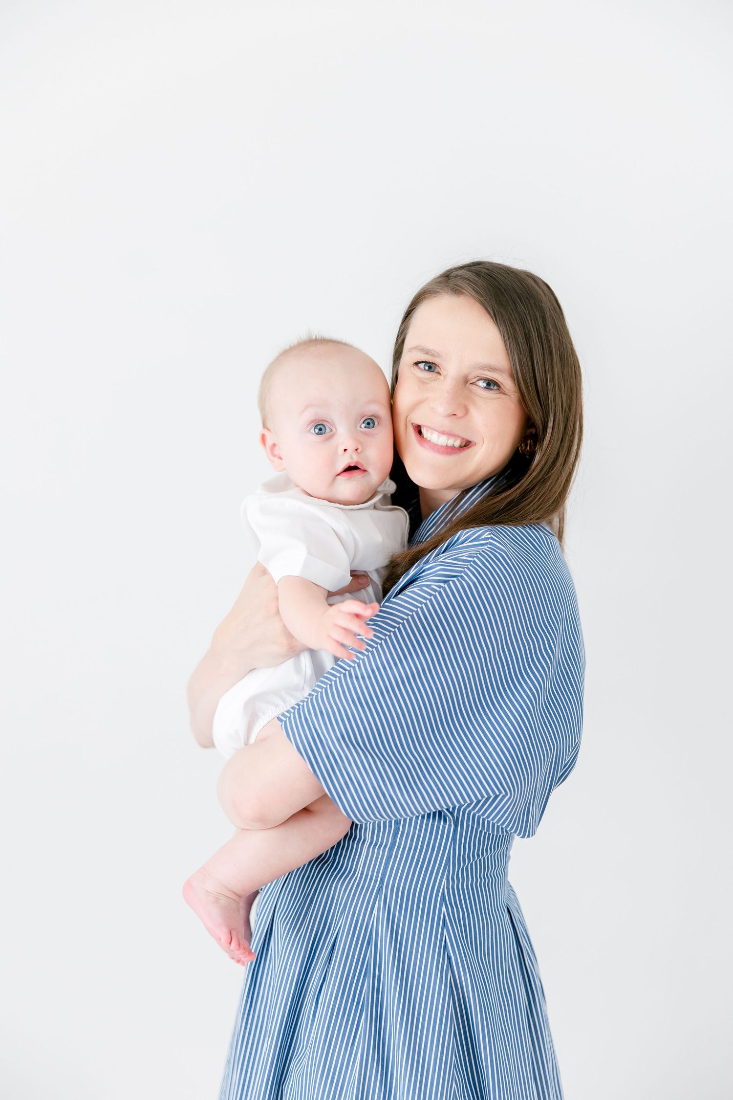 A heartwarming portrait against a white background showing a baby in white being held in a blue striped dress.