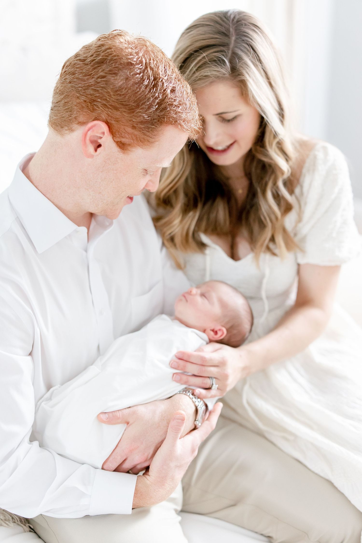 Parents in white outfits share tender moment gazing at their newborn baby in soft natural light.