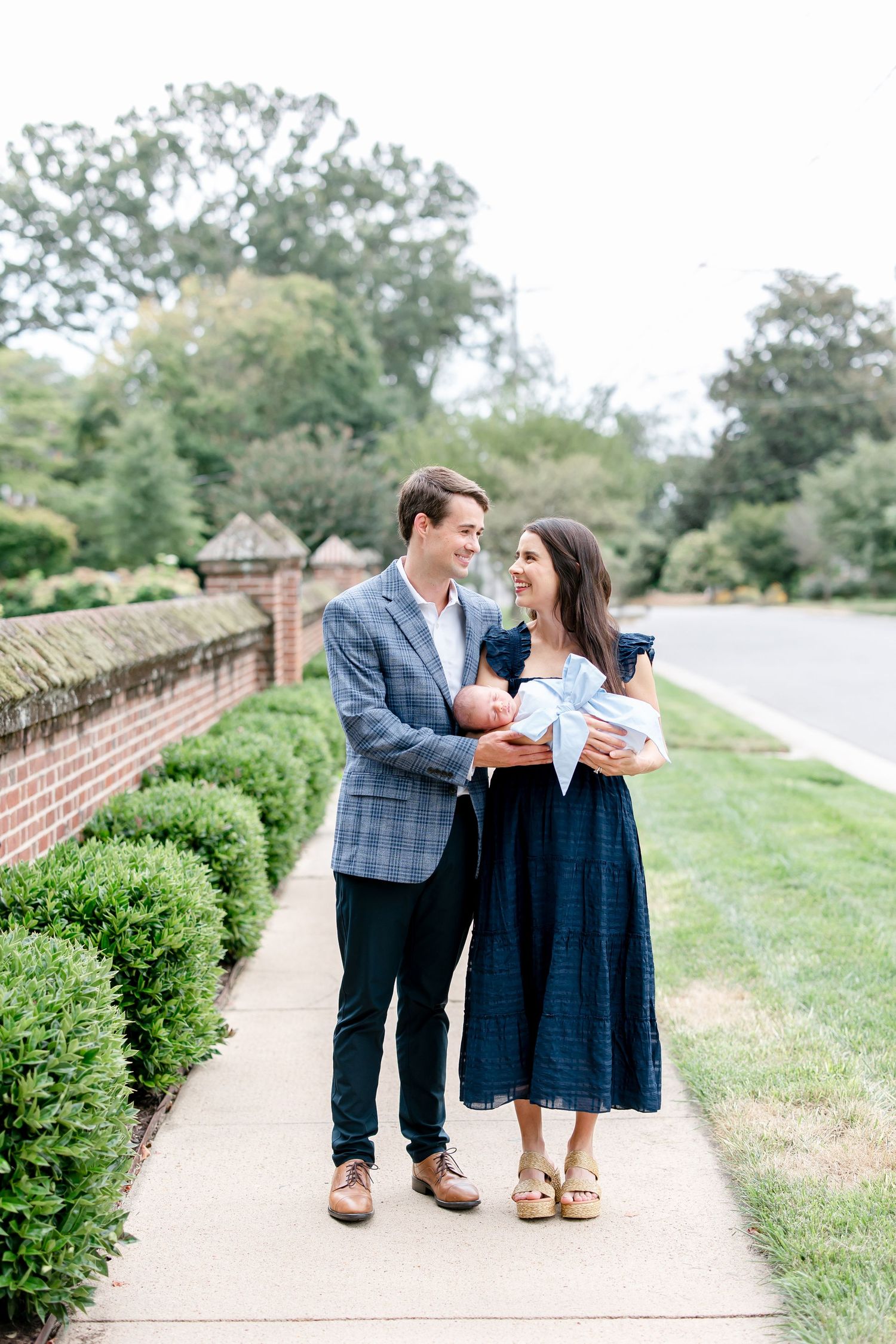 A couple embraces while walking along a brick-lined pathway surrounded by green hedges and trees.