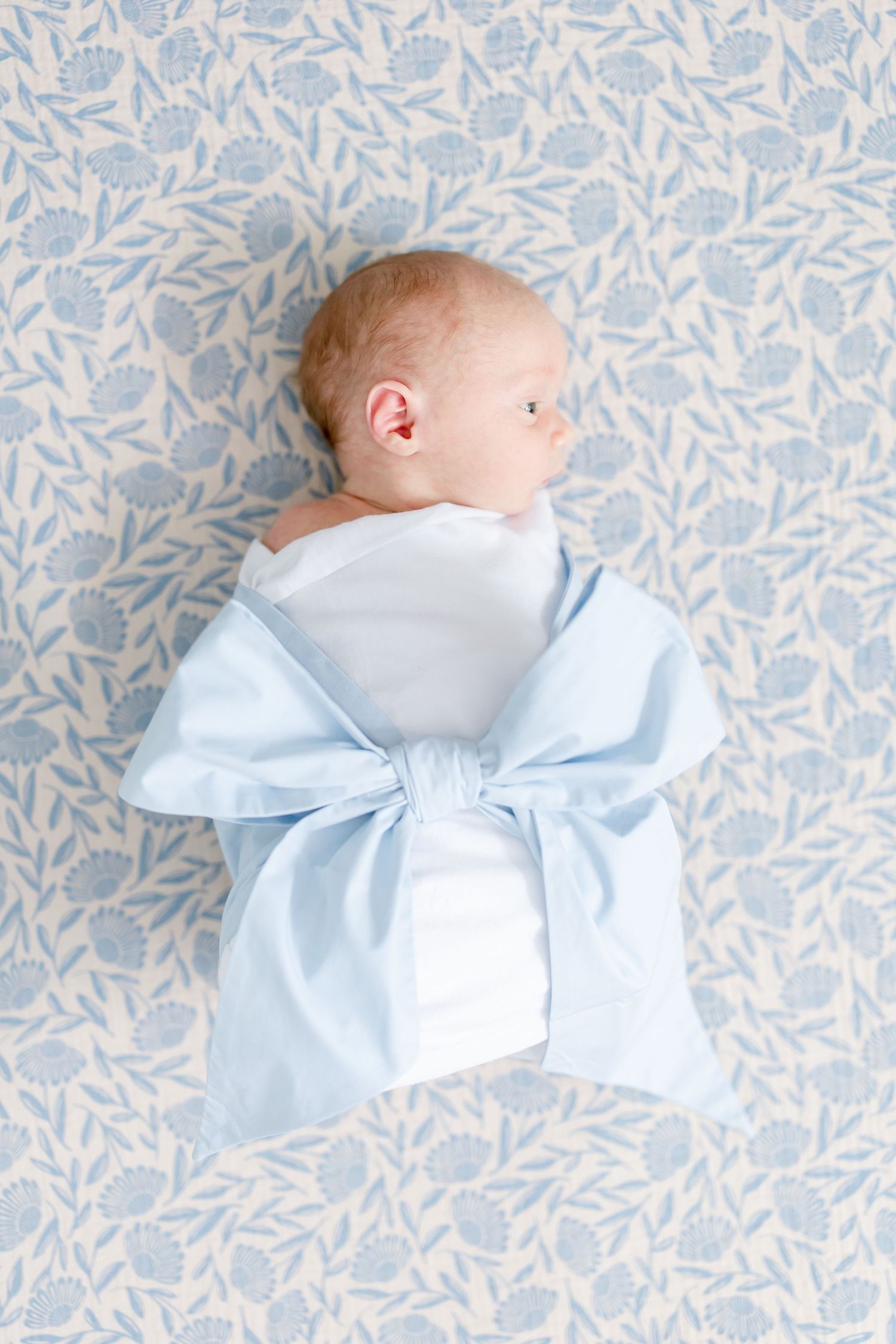 Newborn wearing light blue bow headband rests on delicate floral pattern background.