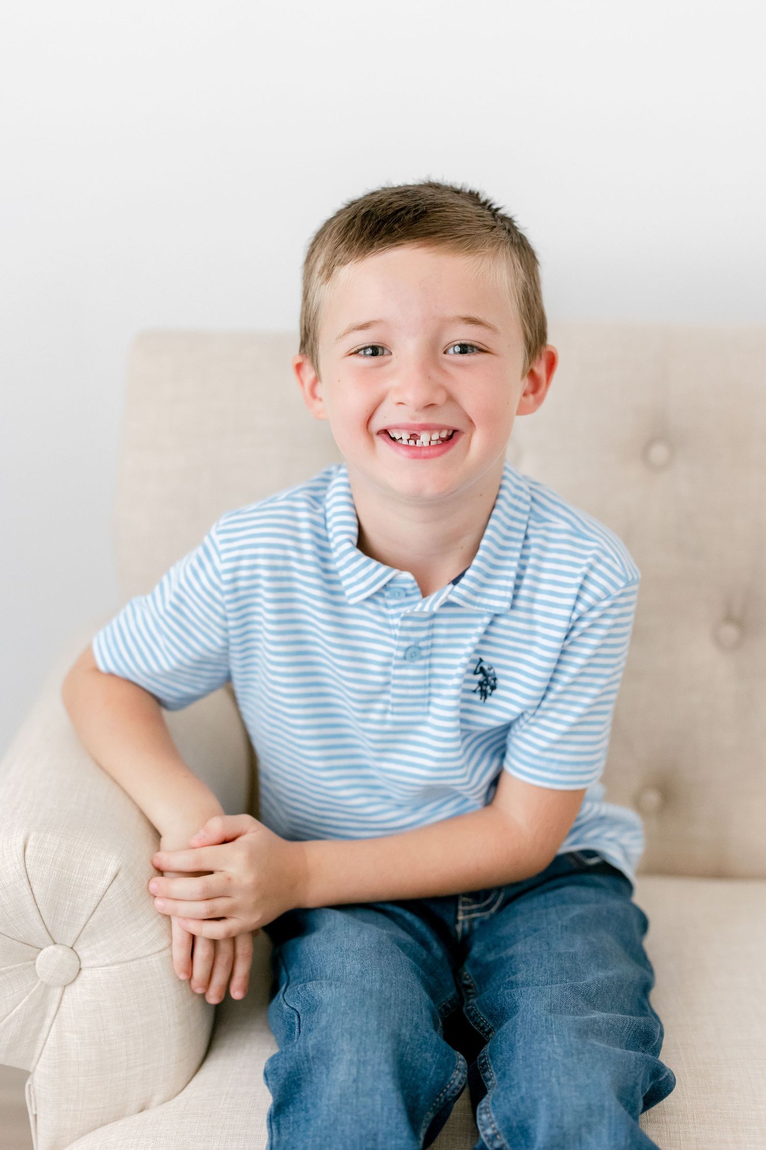 Young child in blue striped polo shirt sitting on cream couch showing joyful expression.