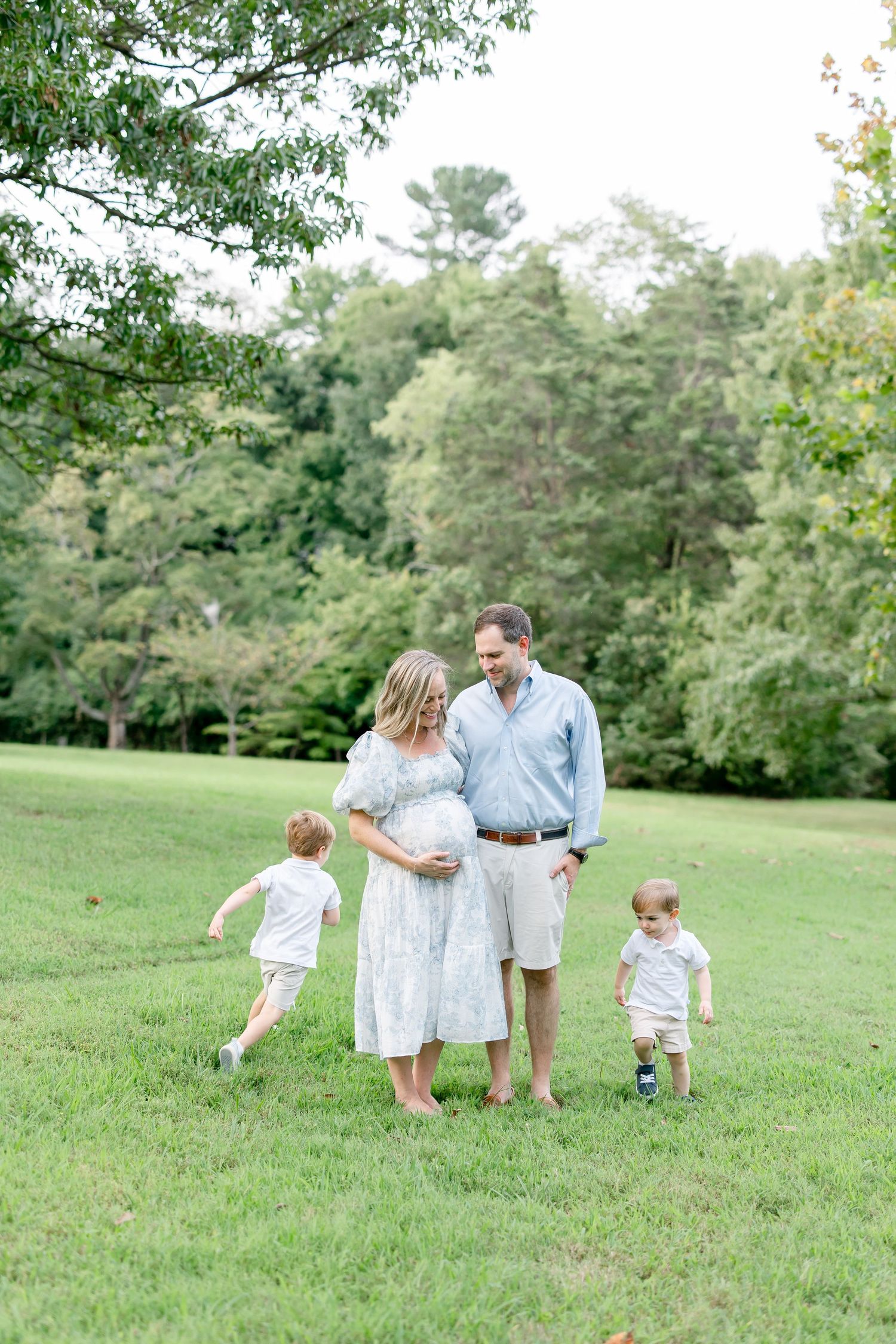 Two children in white outfits walk through a lush green field with their parents on a sunny day.