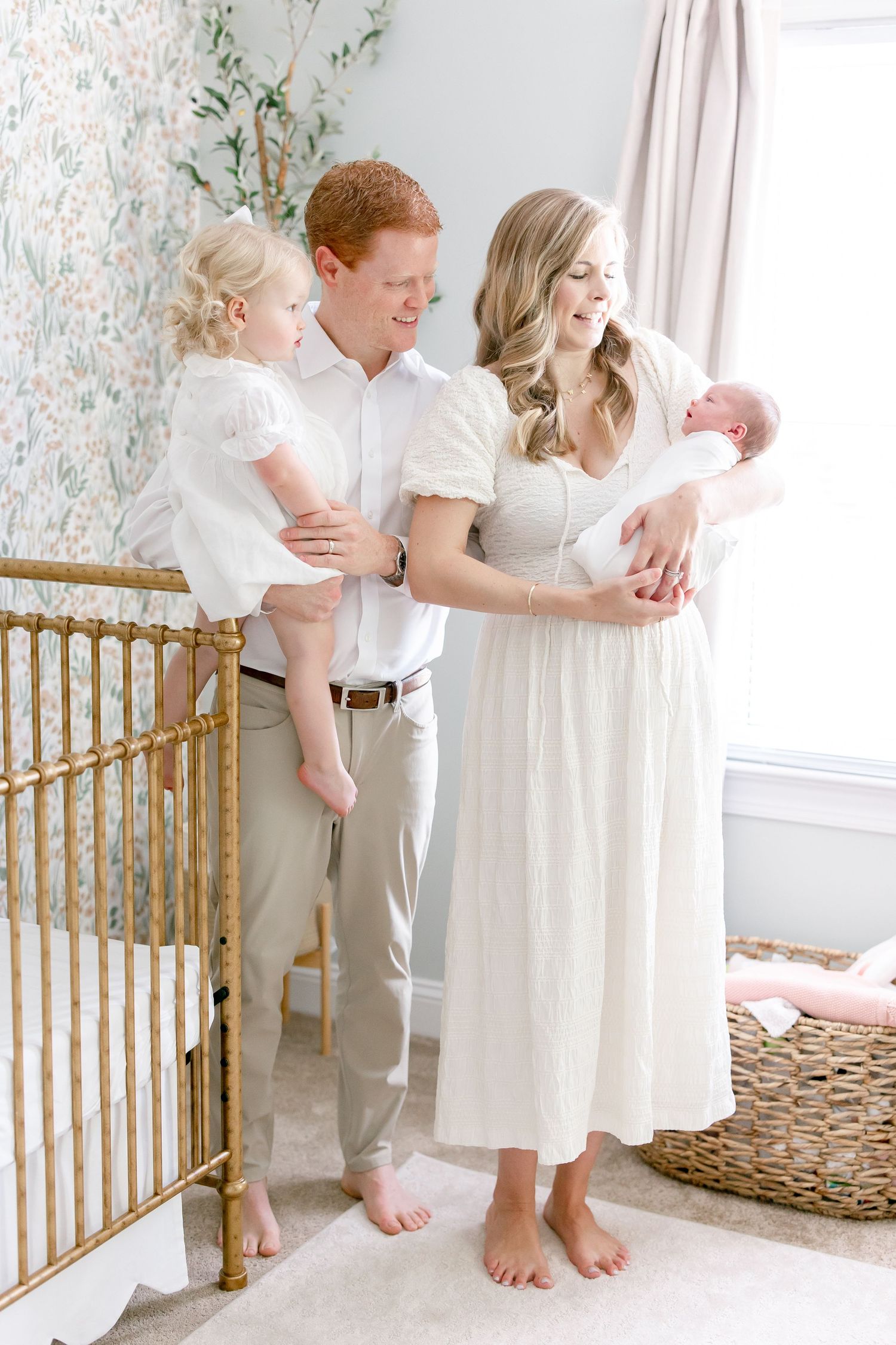 A family of four poses in a nursery with floral wallpaper and a gold crib.