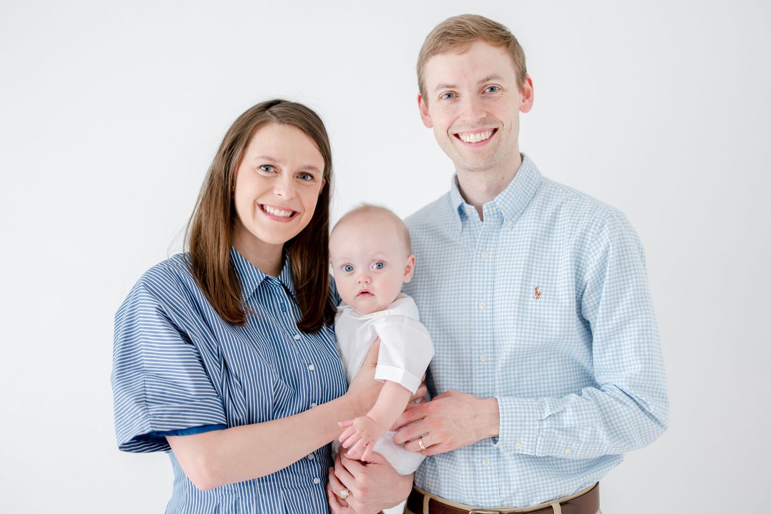 A family in coordinating blue outfits poses together with their baby against a white backdrop.
