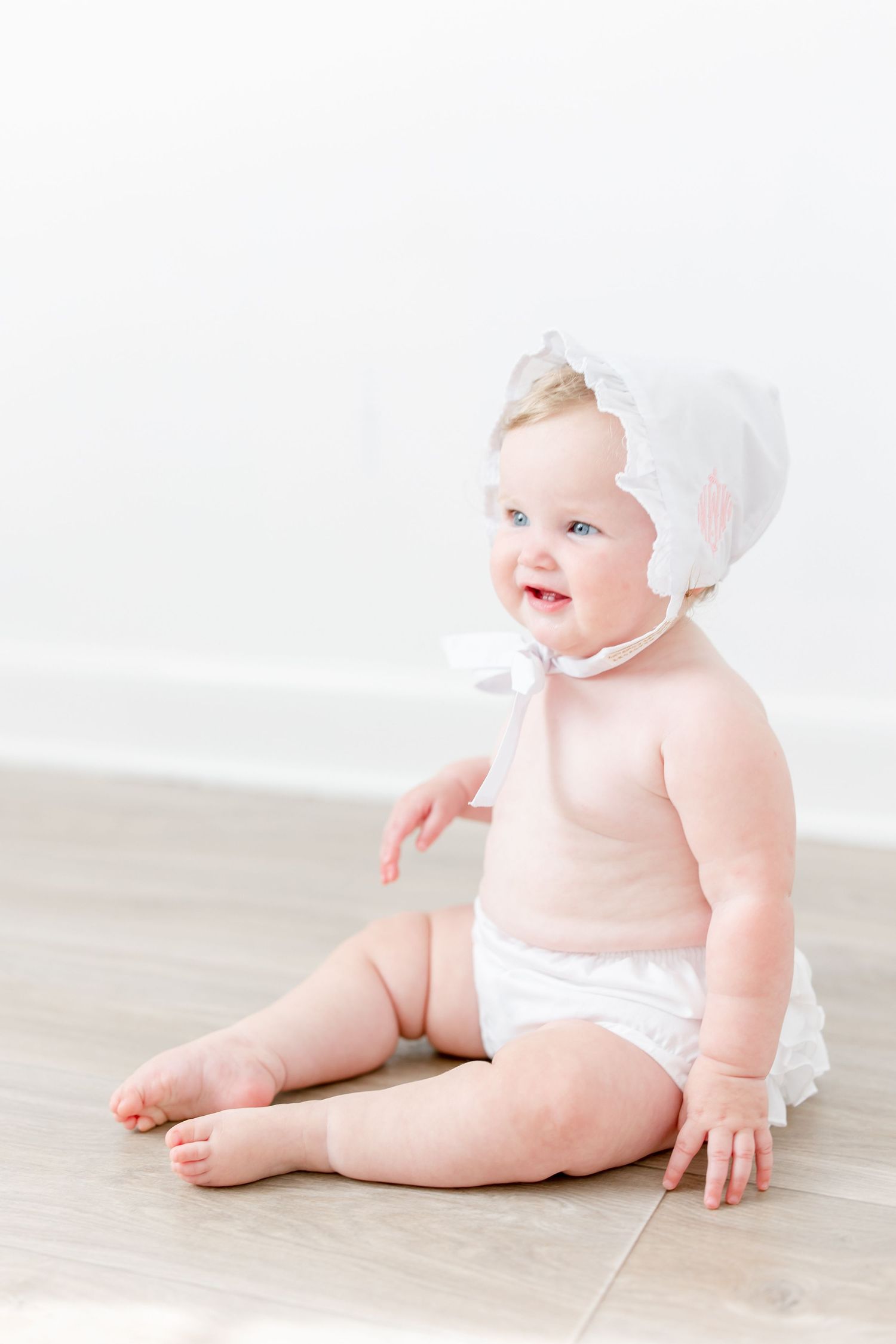 Adorable baby in white bonnet and diaper lying on light wooden floor.