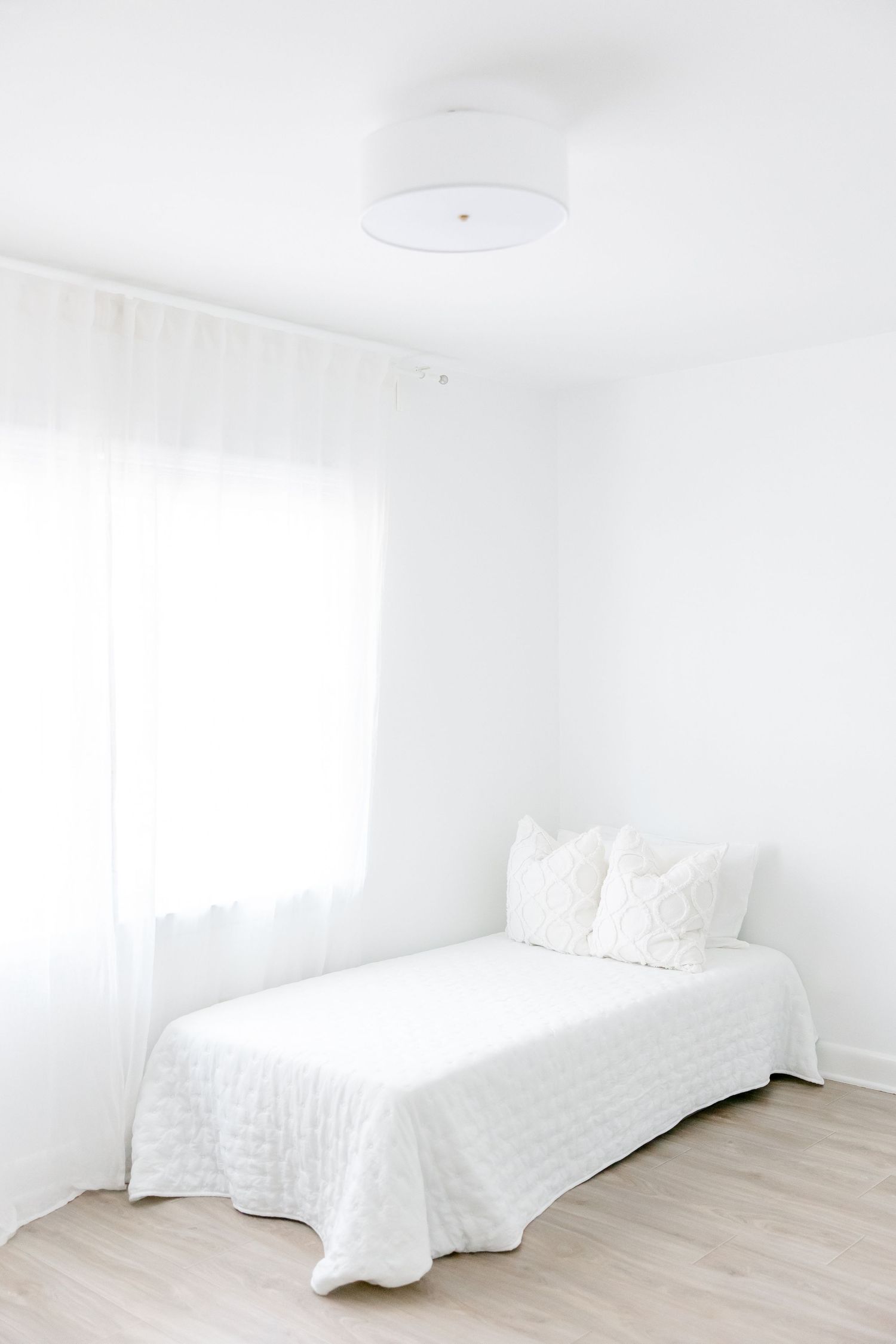 Minimalist white bedroom with single bed, sheer curtains and light wood flooring in a bright, airy space.