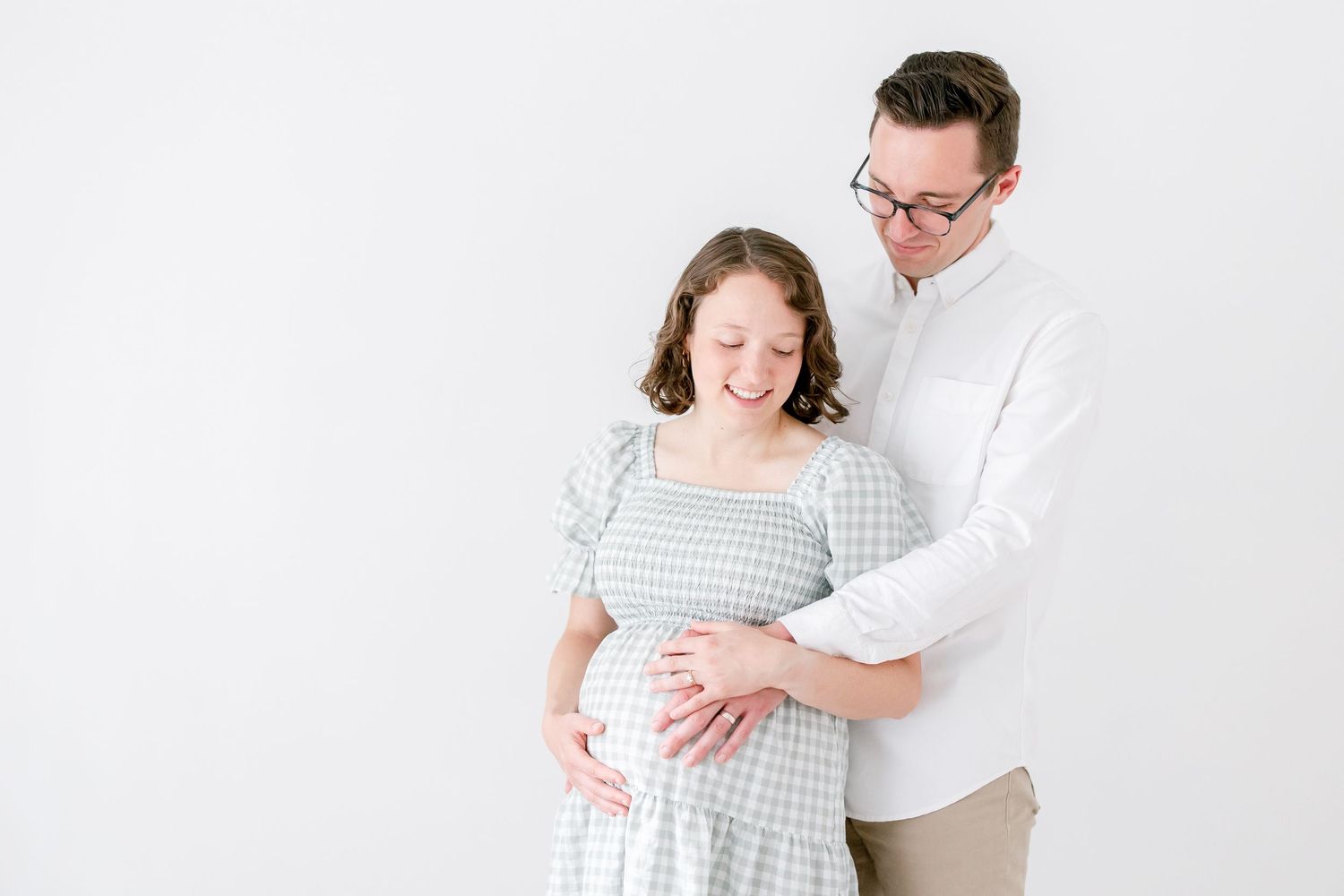 A romantic maternity photo shoot featuring an expectant couple in white and gray outfits against a light background.