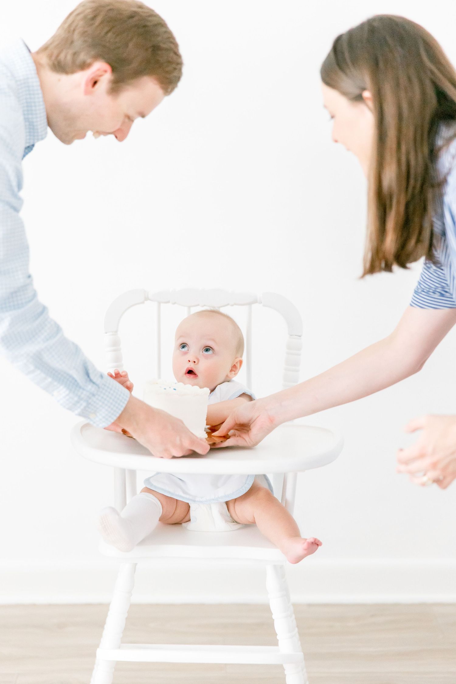 Parents preparing cake smash photo setup with baby in white high chair.