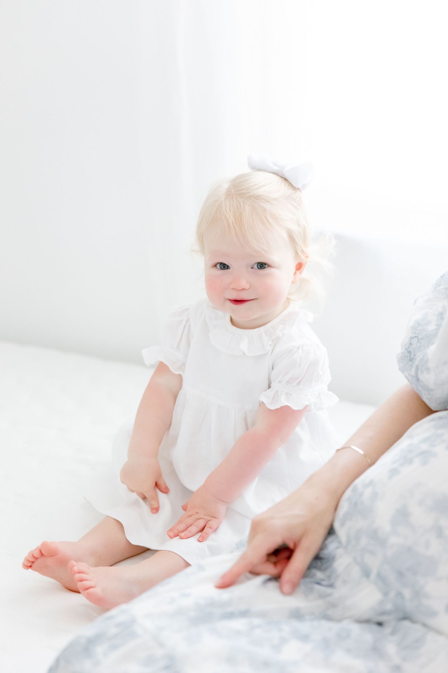 Sweet toddler in white dress sitting on white bedding with delicate details.