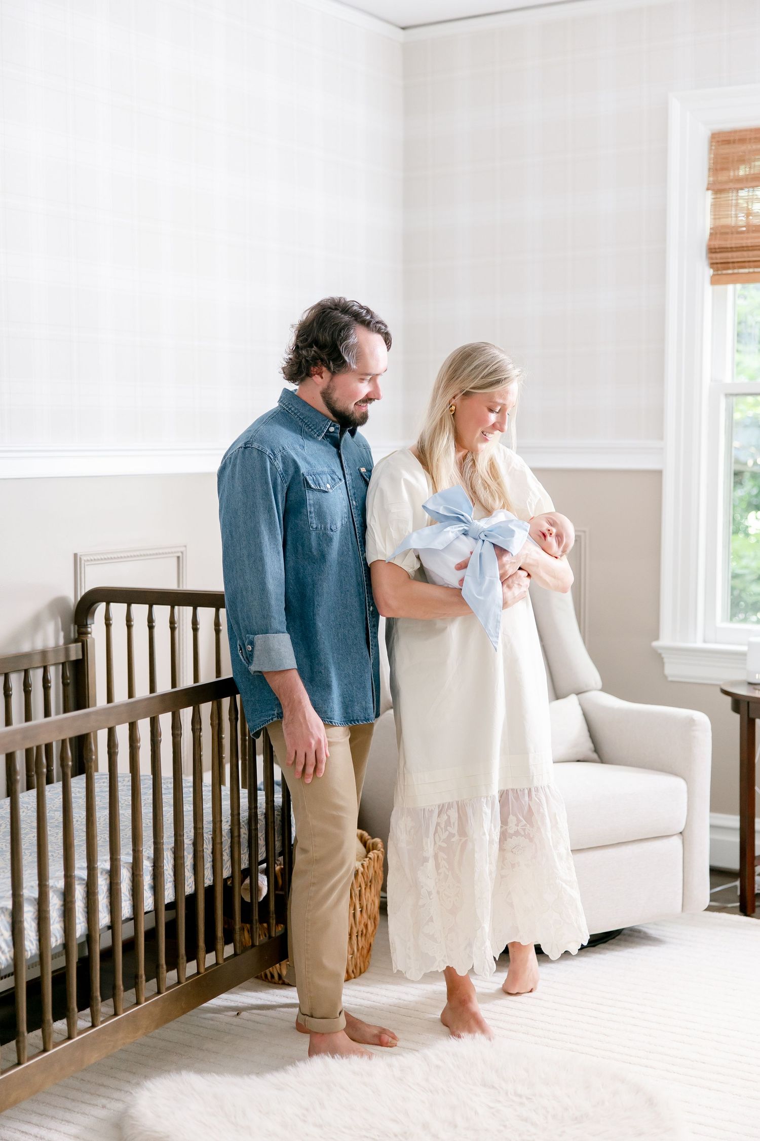 A couple stands with their newborn baby in a bright nursery with white walls and dark wood crib.