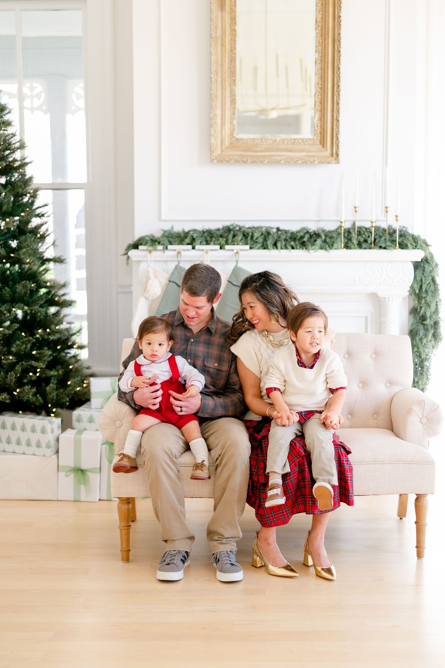 Family of four sitting on a cream sofa by a decorated Christmas tree and fireplace, wearing festive red and plaid outfits.