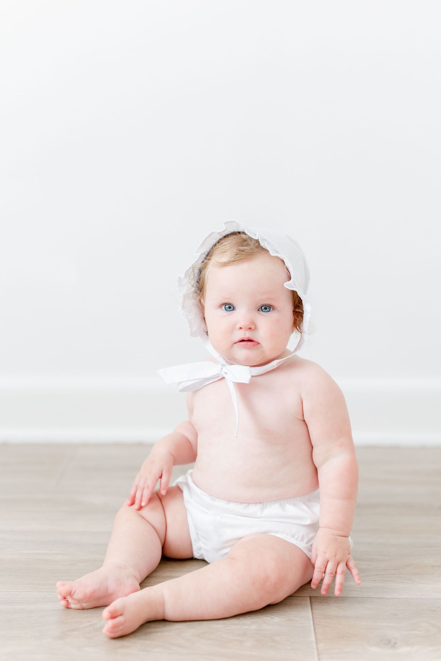 Baby wearing a pink top and white diaper sits on light hardwood floor against white wall.