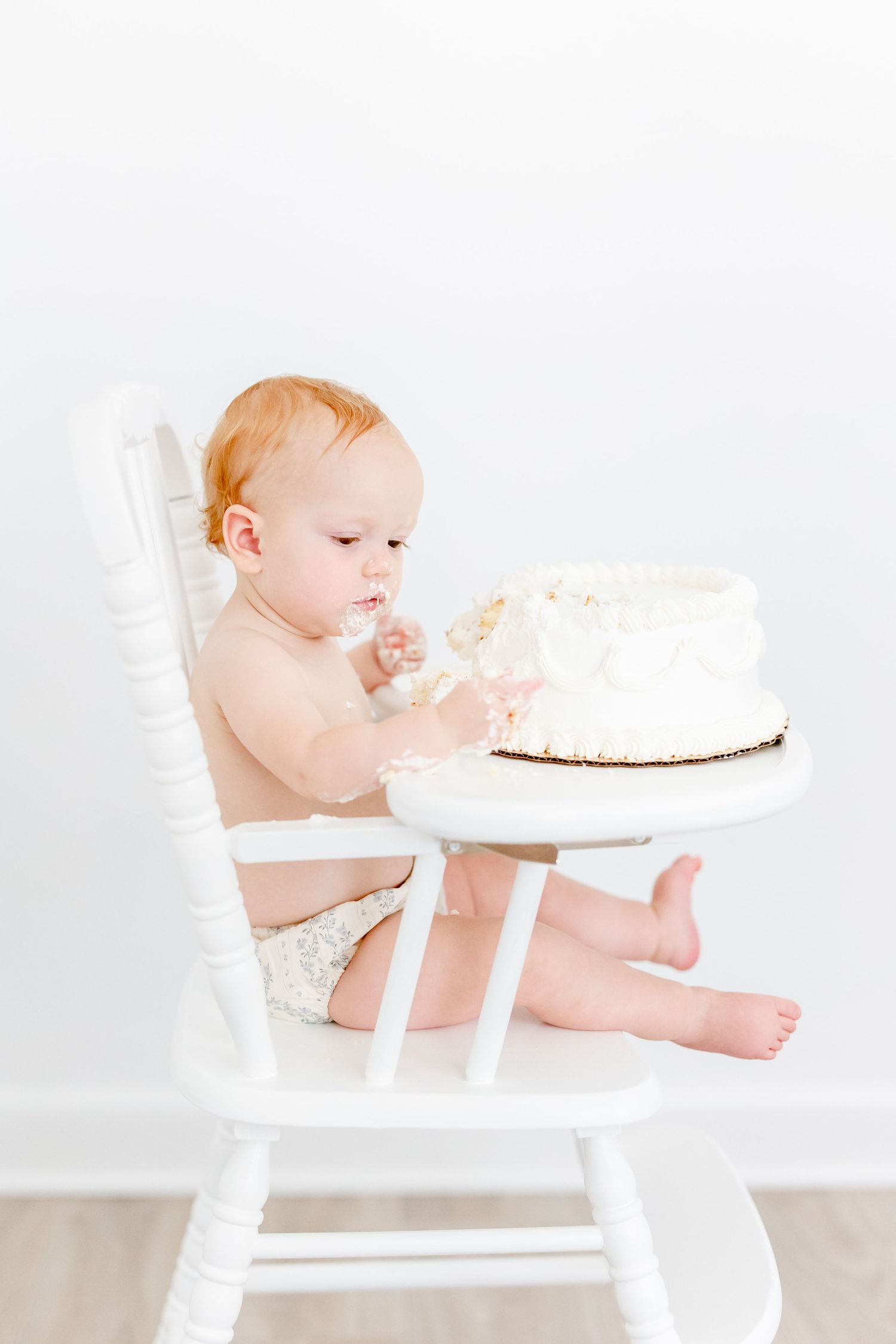 Baby in white high chair reaching for white cake during cake smash photo session against neutral background.