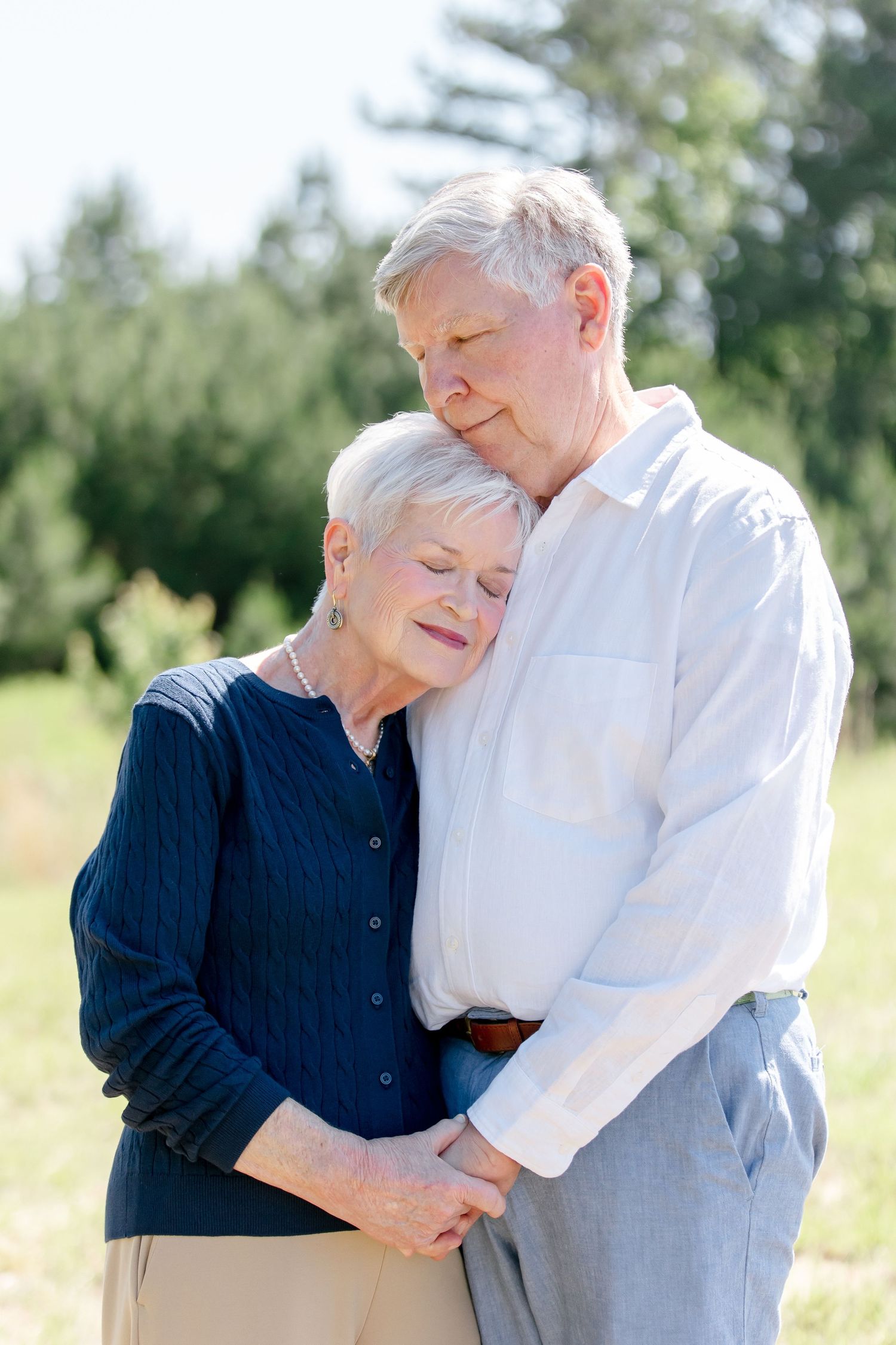 A tender outdoor moment between an elderly couple as they embrace in casual attire against a sunny background.