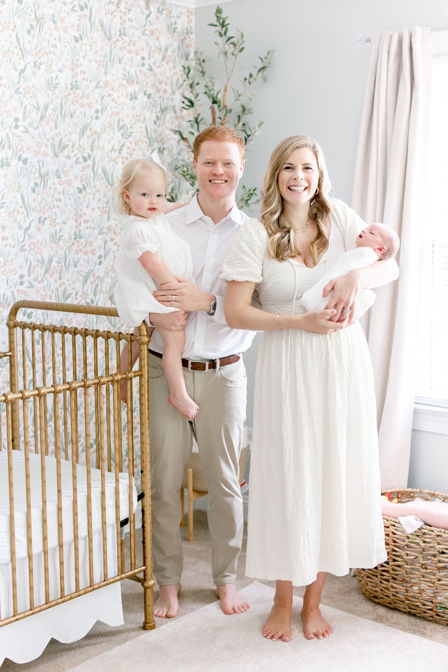 Young family in white outfits stands barefoot in nursery room with gold crib and floral wallpaper.