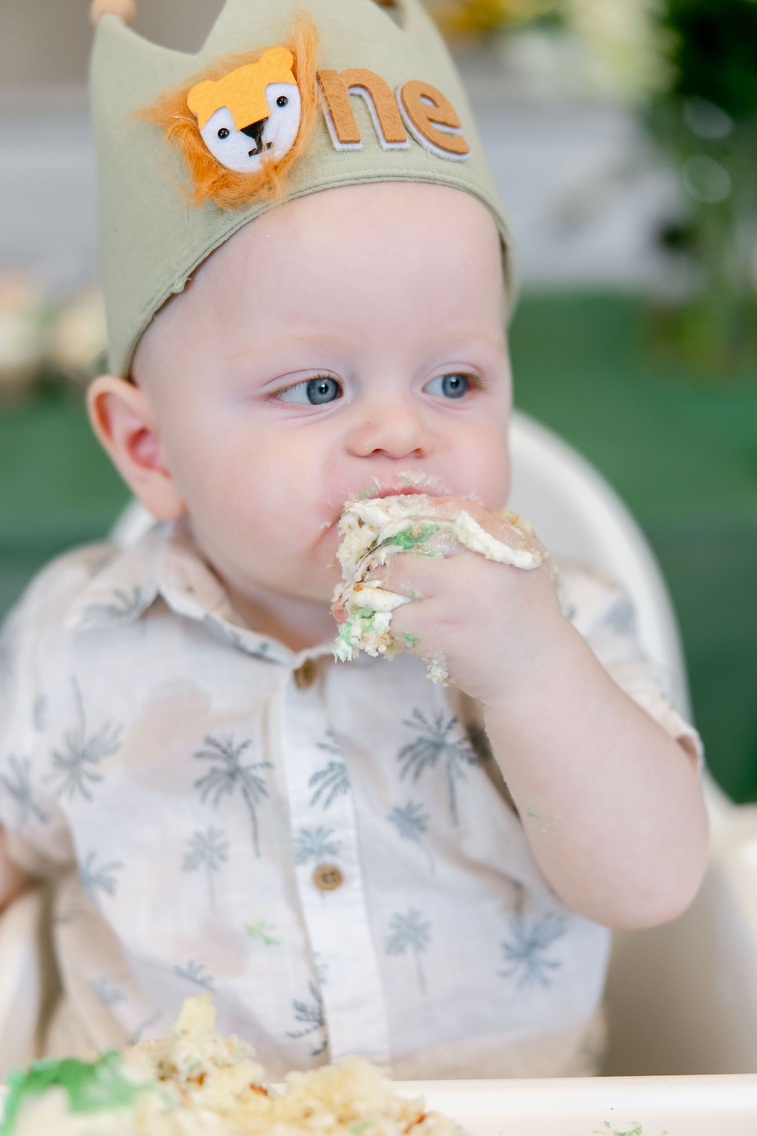 Baby wearing lion crown eating cake messily during first birthday celebration.