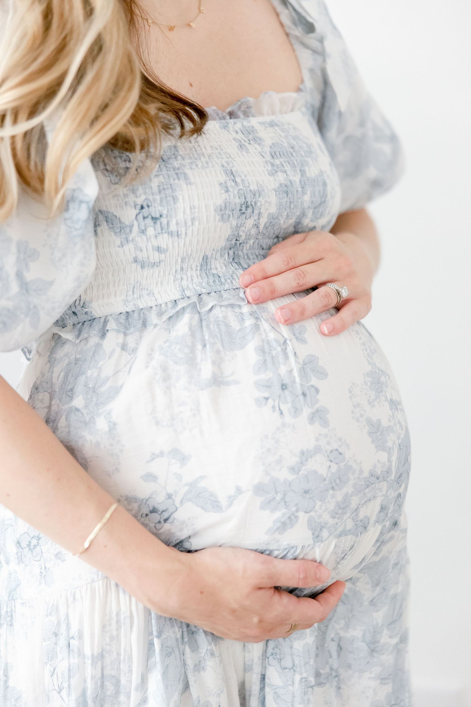 A pregnant woman in a soft blue and white floral dress gently cradles her baby bump against a light background.