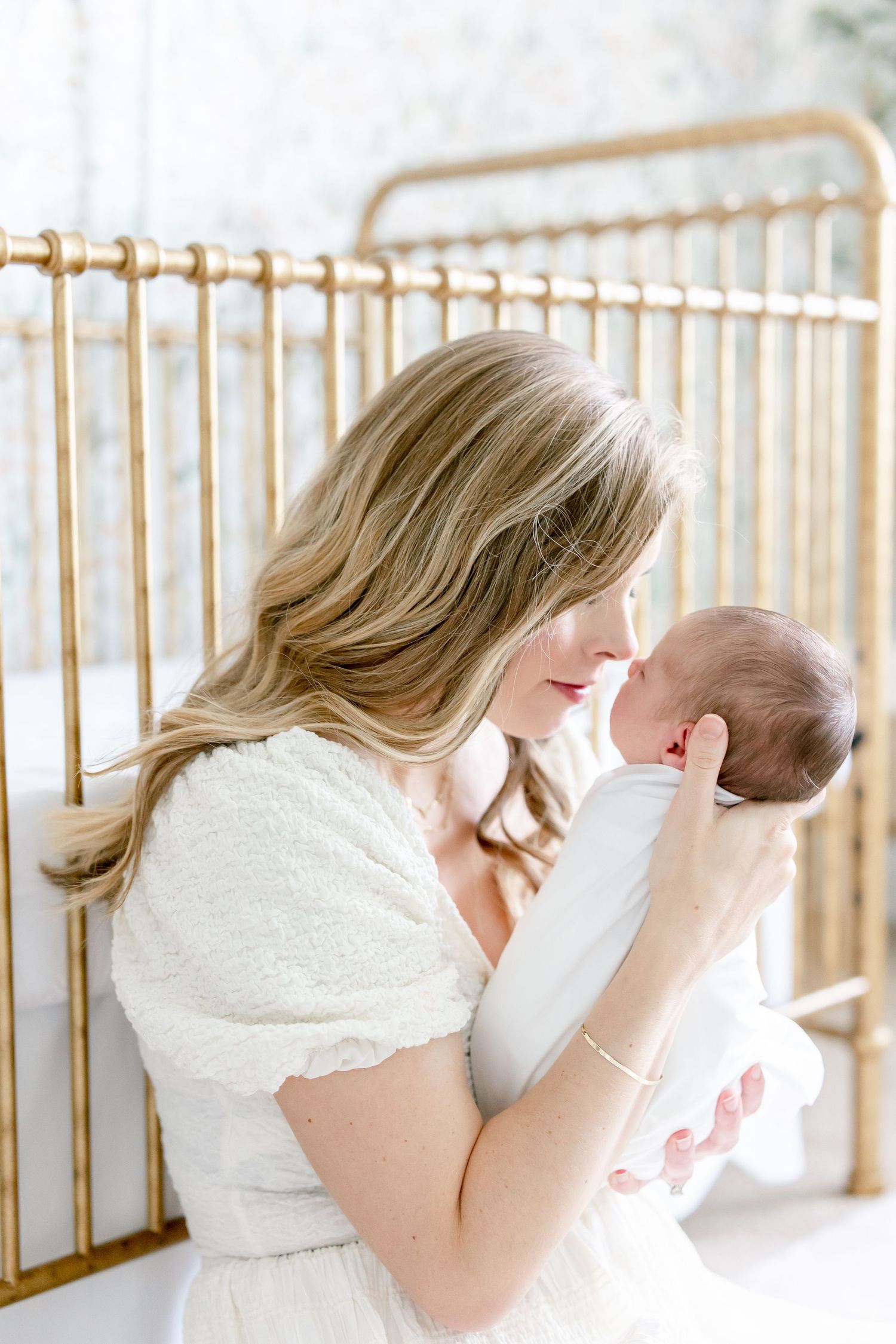 Mother in white dress holds newborn near gold crib in soft natural light.