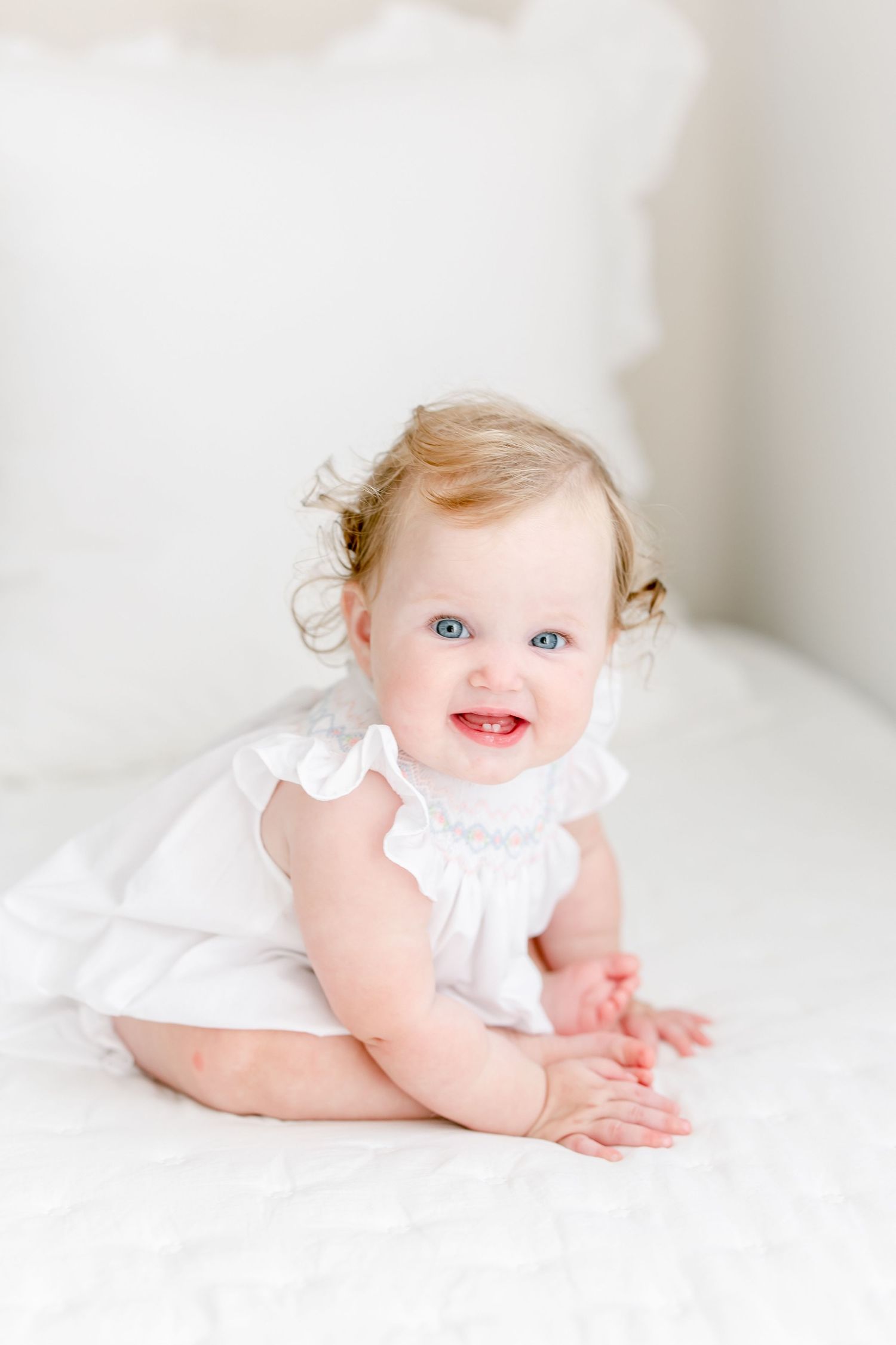 Baby in white ruffled dress sits on light colored bedding while smiling and playing.