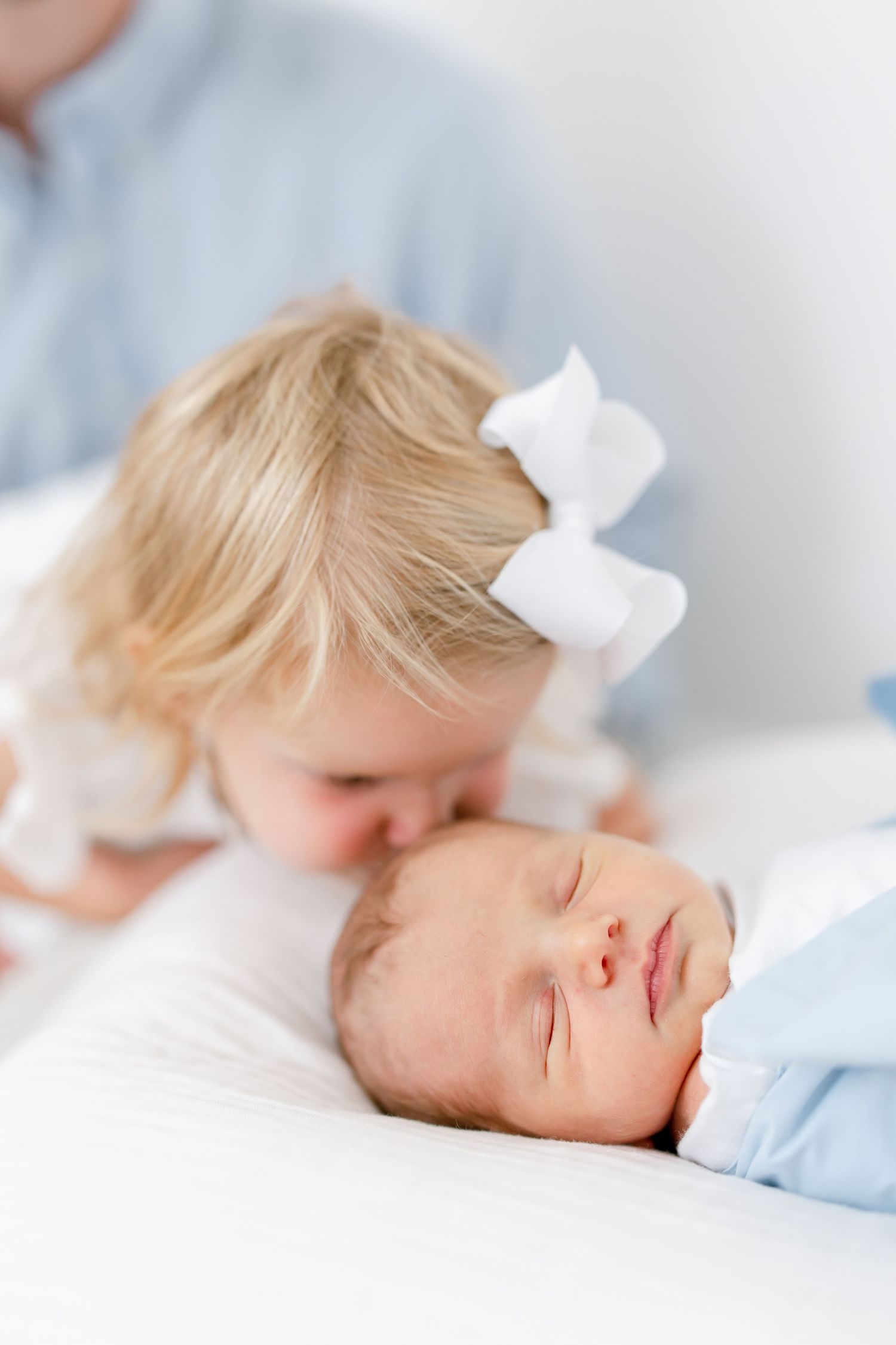 A gentle moment captured as a child with a white bow leans over a sleeping baby in soft light.