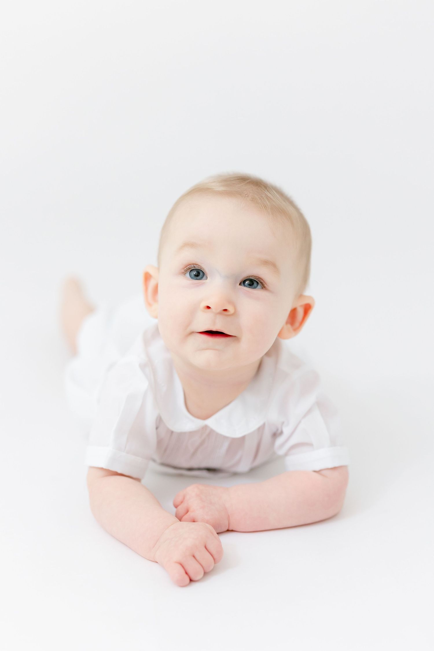 Smiling baby with blue eyes lying on their tummy in a white outfit against a clean white studio background.